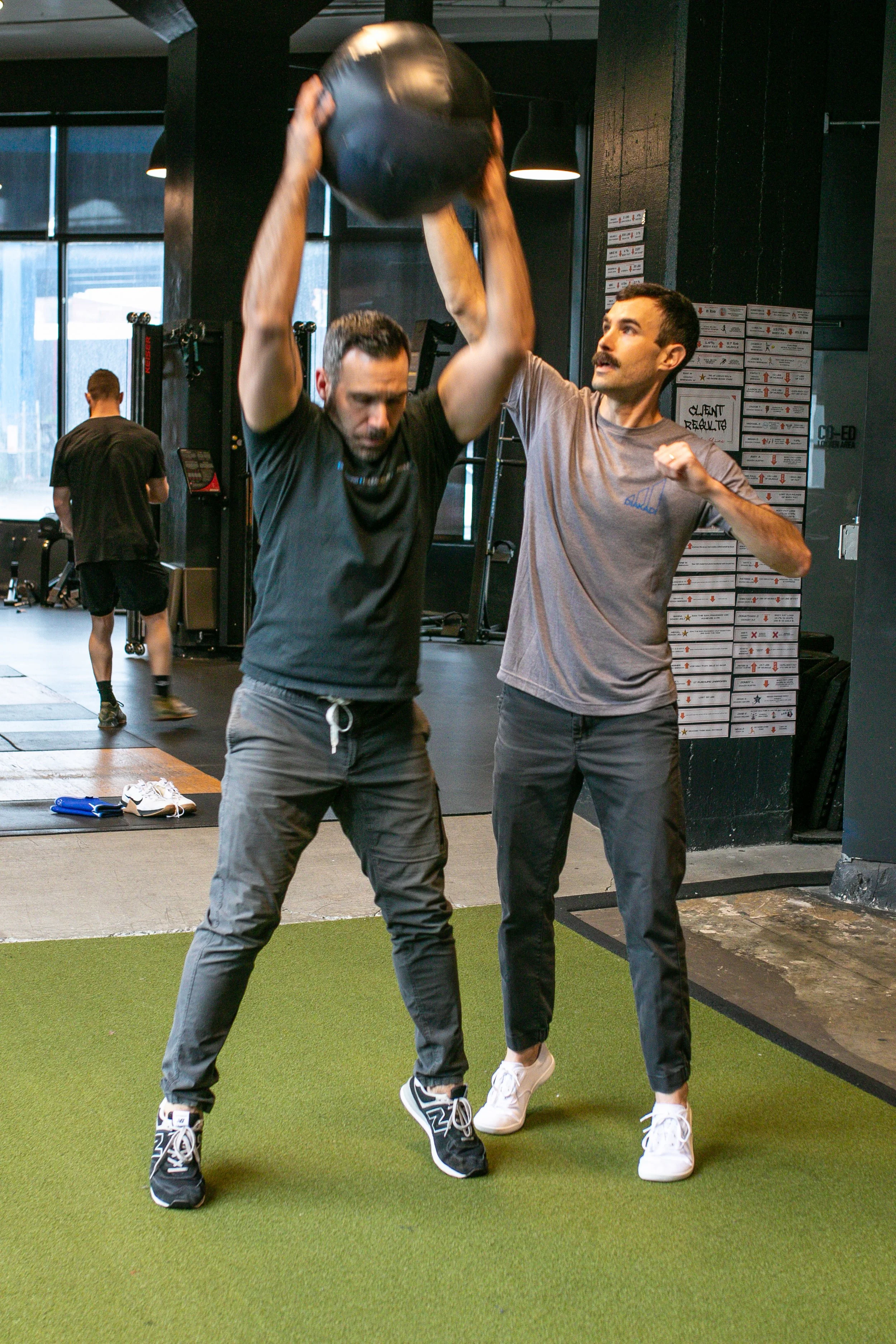 A man lifting a medicine ball overhead with assistance from a trainer in a gym.