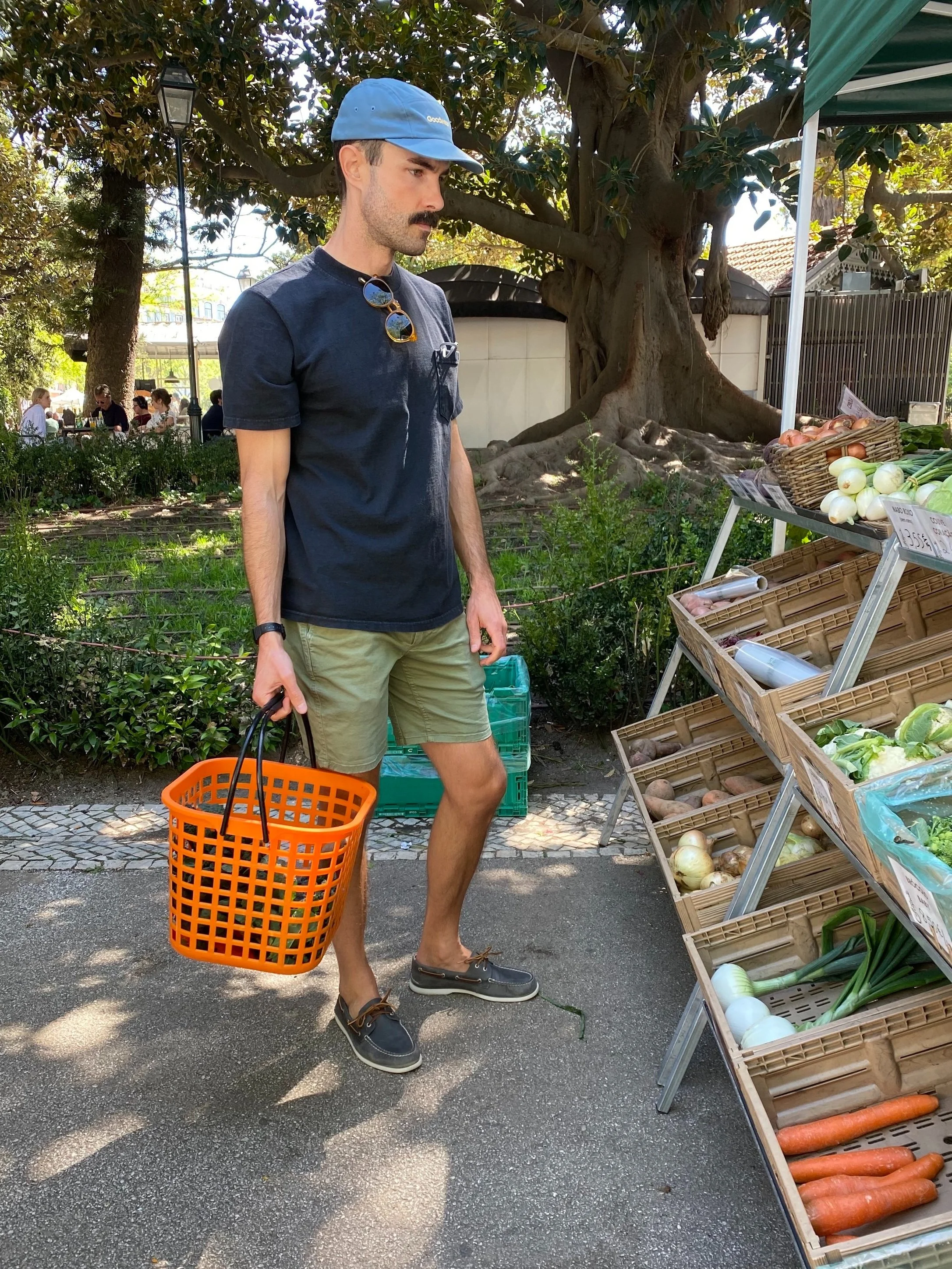 A man shopping at an outdoor farmers market, carrying an orange basket, looking at vegetables like onions, carrots, and green onions displayed in wooden bins.