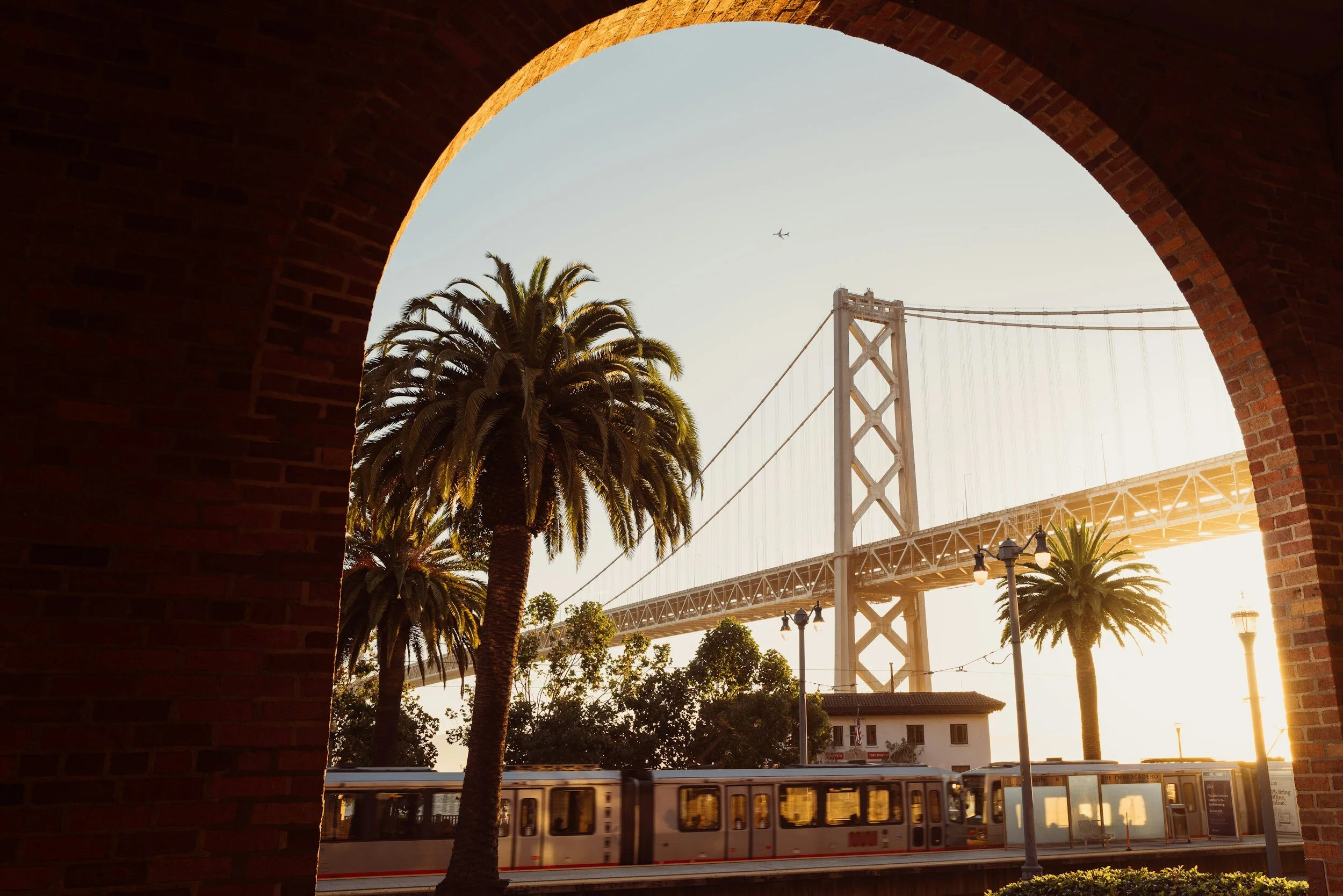 View of the San Francisco Bay Bridge and a train passing by, framed by a brick archway with palm trees in the foreground during sunset.