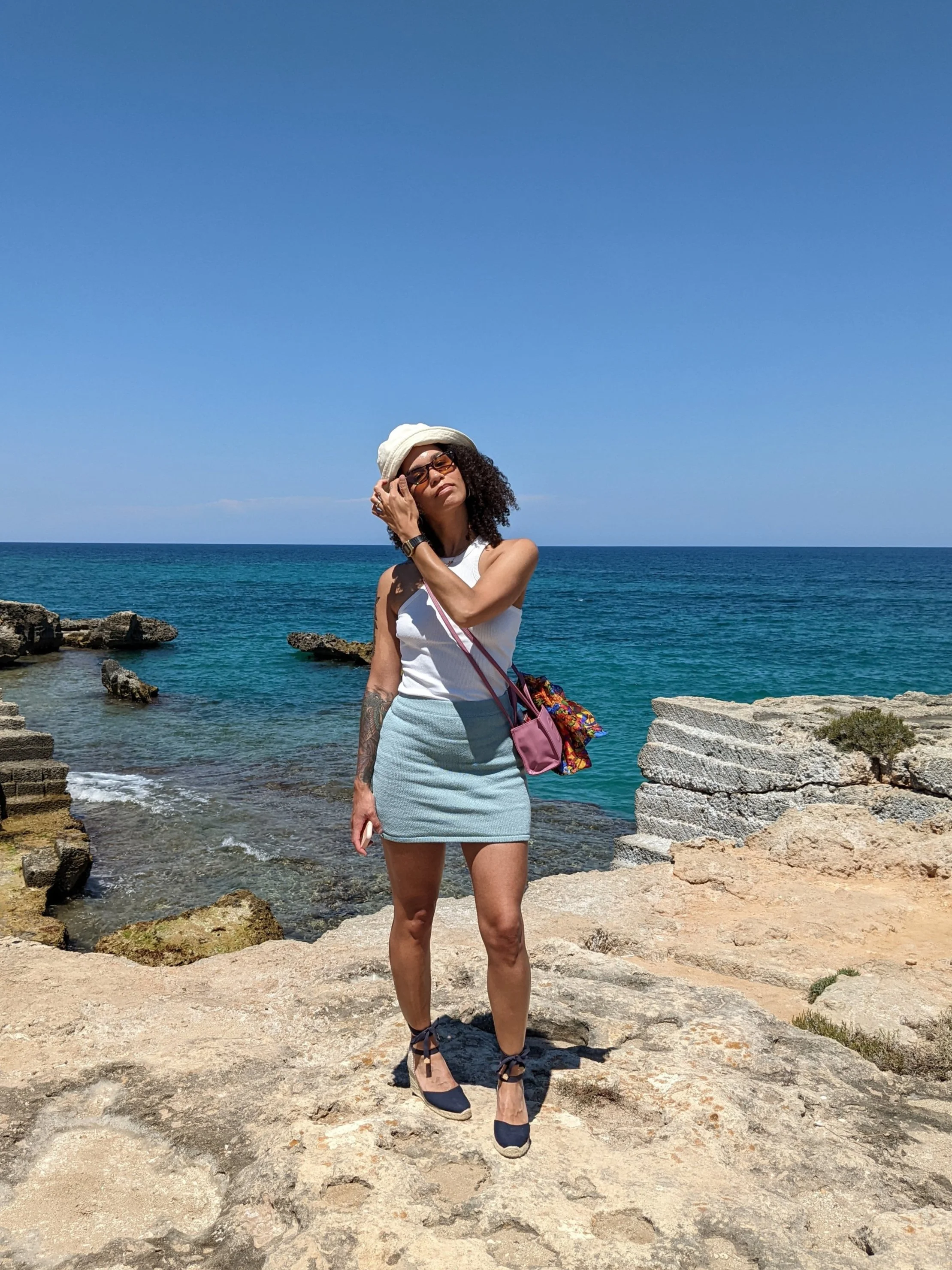 Woman with curly hair, wearing sunglasses, a hat, a white tank top, and a light blue skirt, posing by the rocky shore with the ocean and clear blue sky in background.