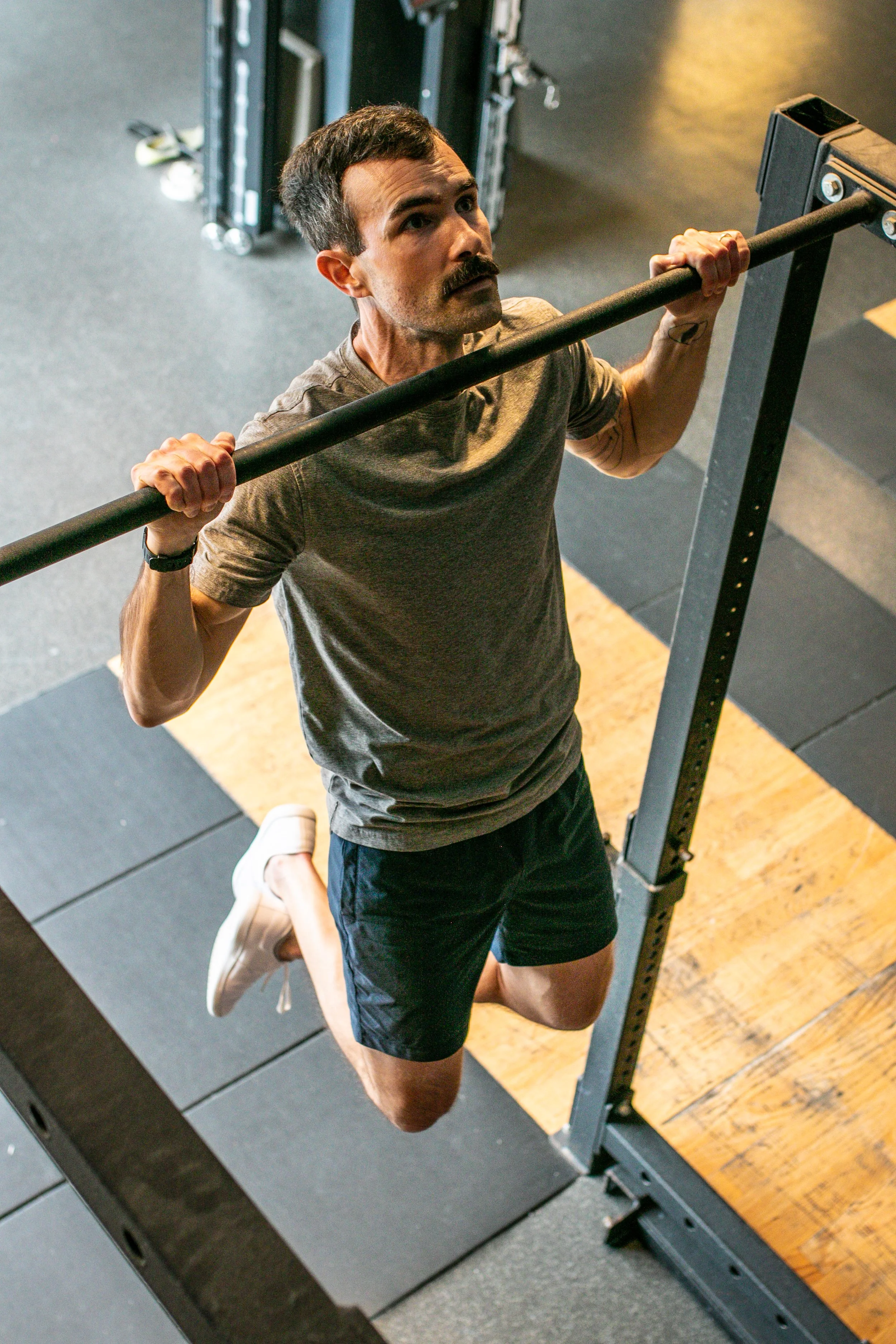 A man performing a pull-up exercise in a gym, holding onto a pull-up bar with both hands, over gym mats.