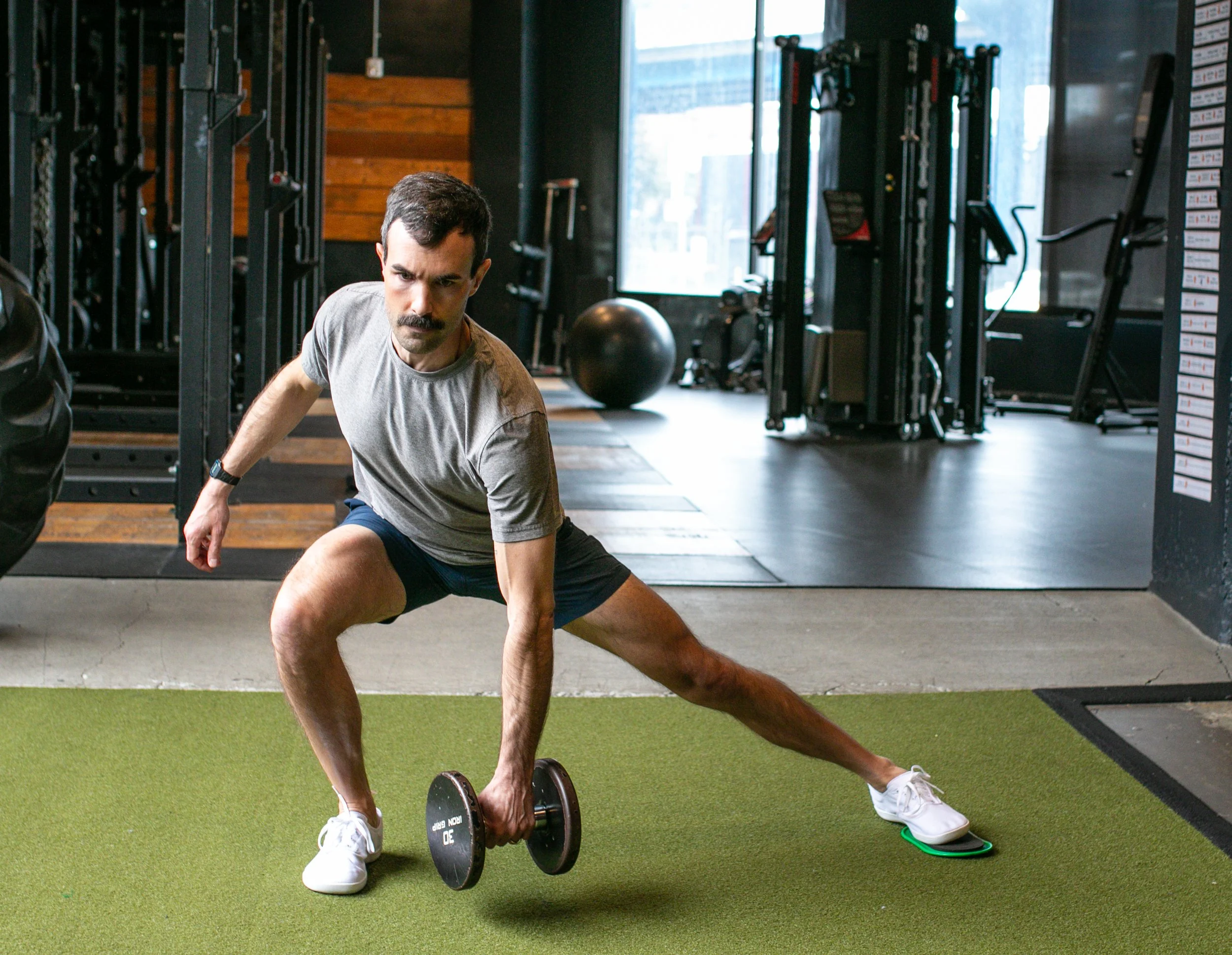 A man in a gym performing a side lunge exercise with a dumbbell in his right hand.