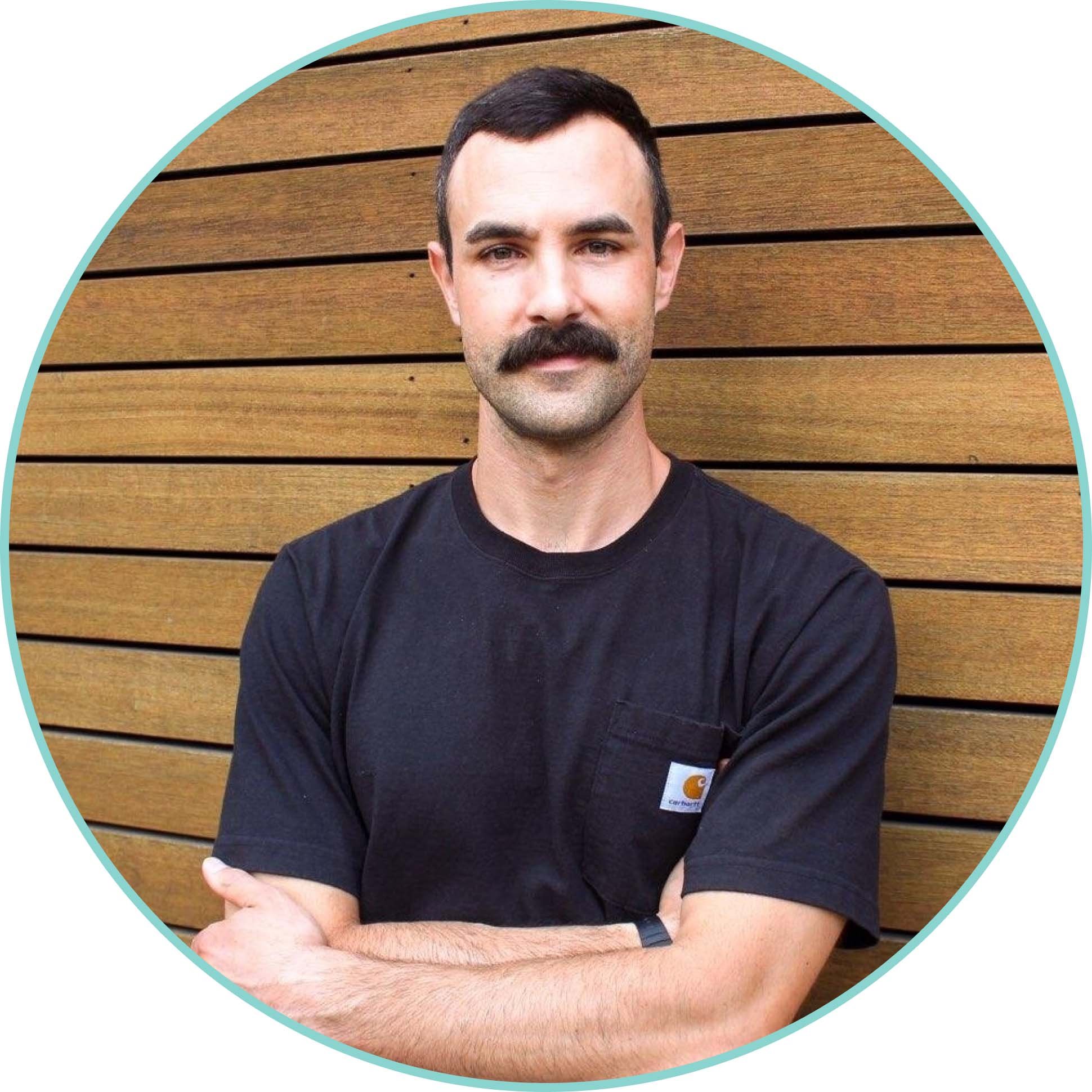 Photo of Dustin Millhollen, a white man, wearing a black t-shirt, with his arms crossed in front of him.