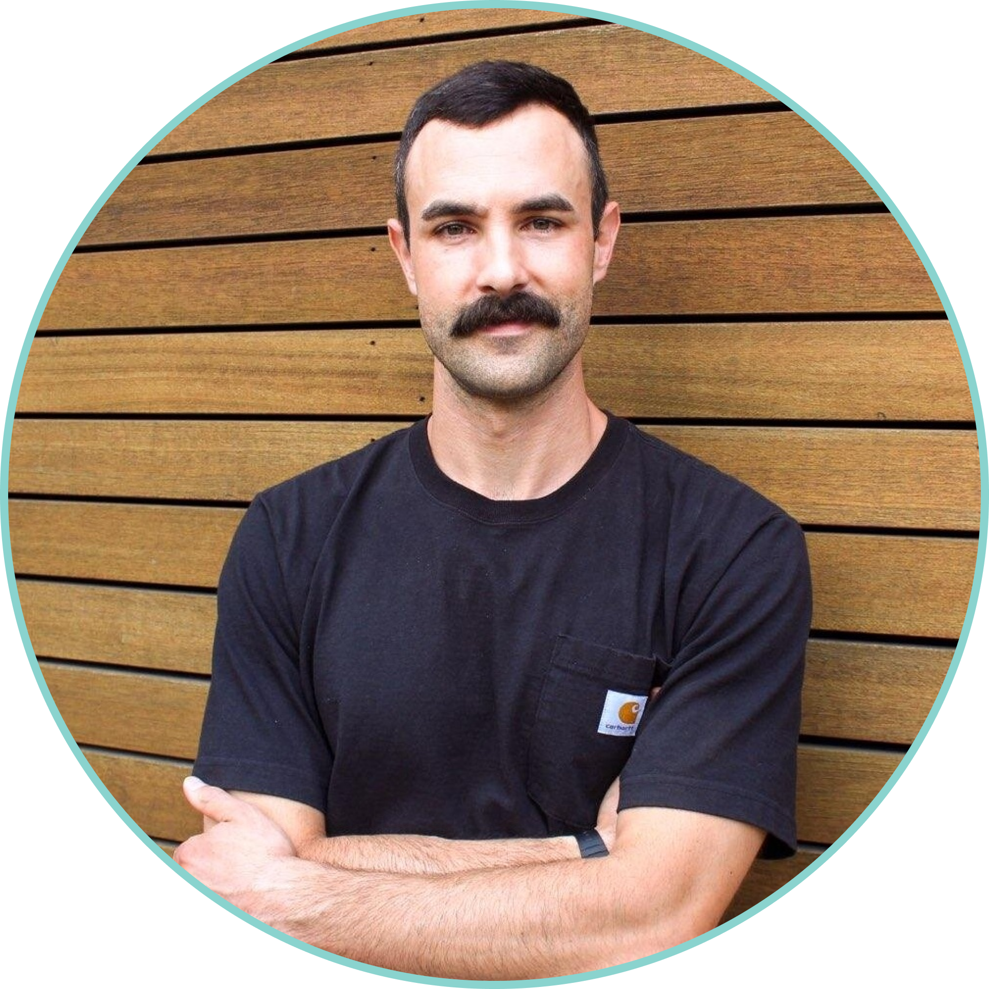 A man with dark hair and a moustache standing with arms crossed in front of a wooden wall, wearing a black Carhartt t-shirt.