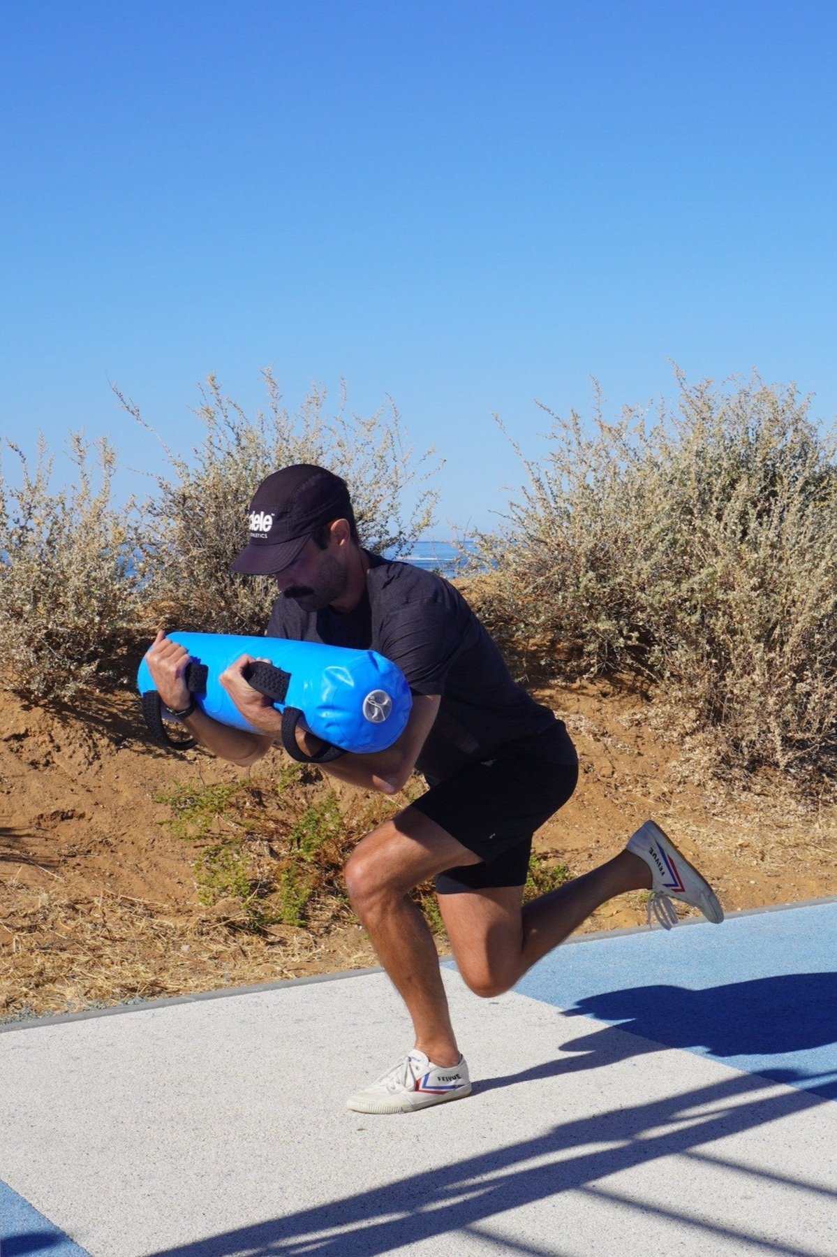 A man running on a trail next to bushes, carrying a blue water bag under his arm, wearing a black cap, black shirt, black shorts, and white sneakers, with a clear blue sky overhead.