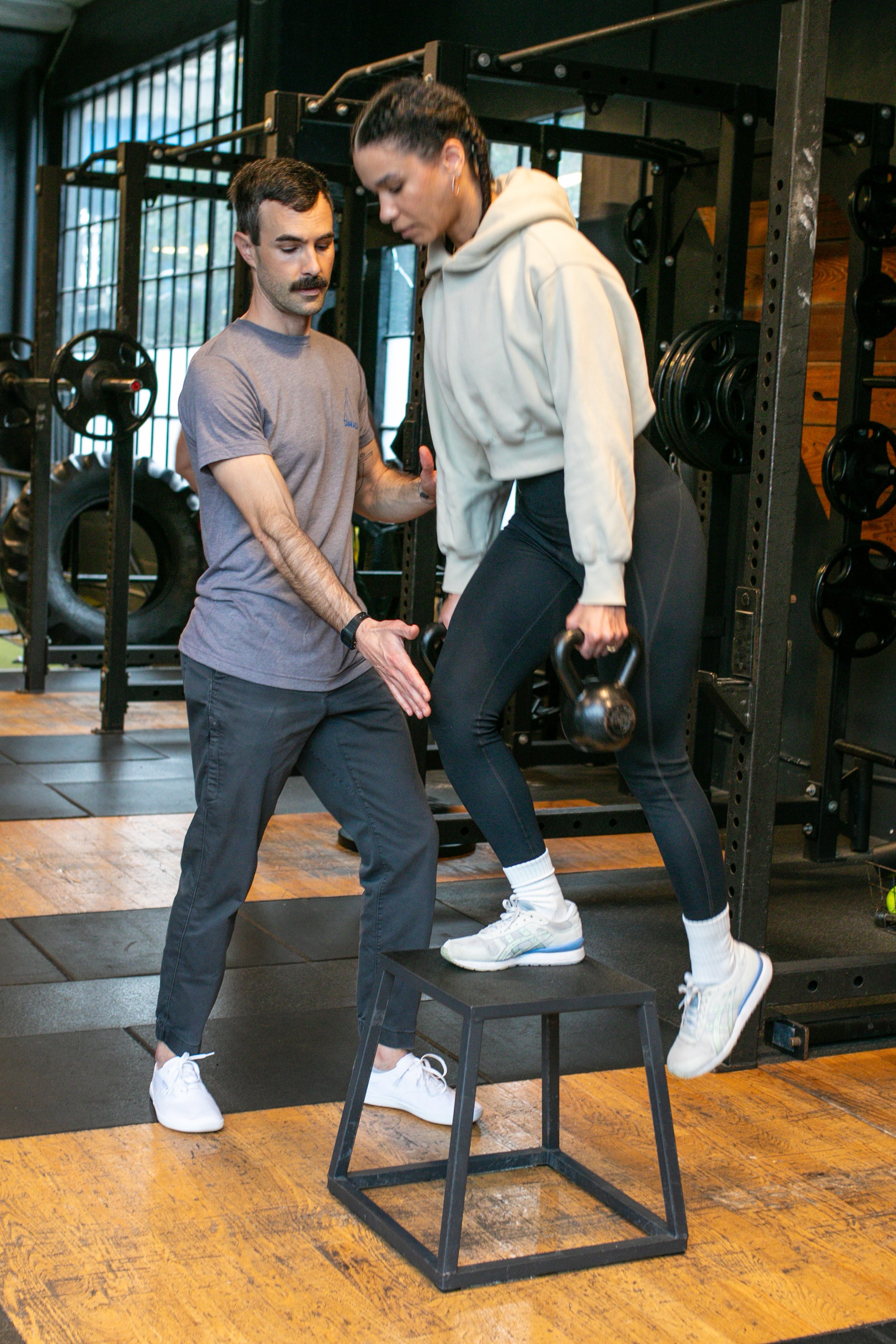 Personal trainer assisting a woman with a kettlebell exercise on a step stool in a gym.