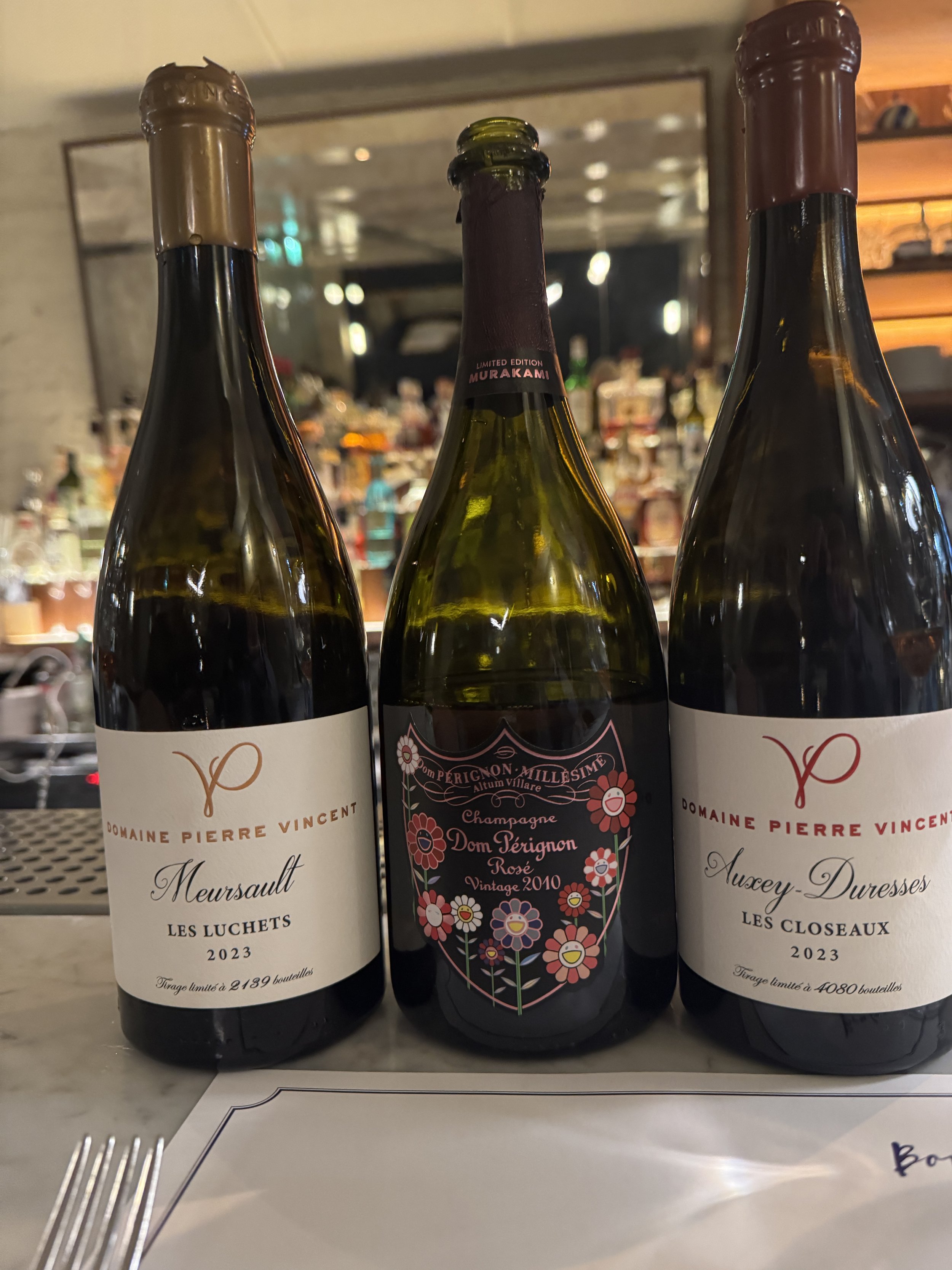 Three wine bottles on a table, with a blurred restaurant background. The bottles are labeled Domaine Pierre Vincent, with the two on the sides being from 2023, and the middle one a vintage 2010 Dom Pérignon Rosé champagne.