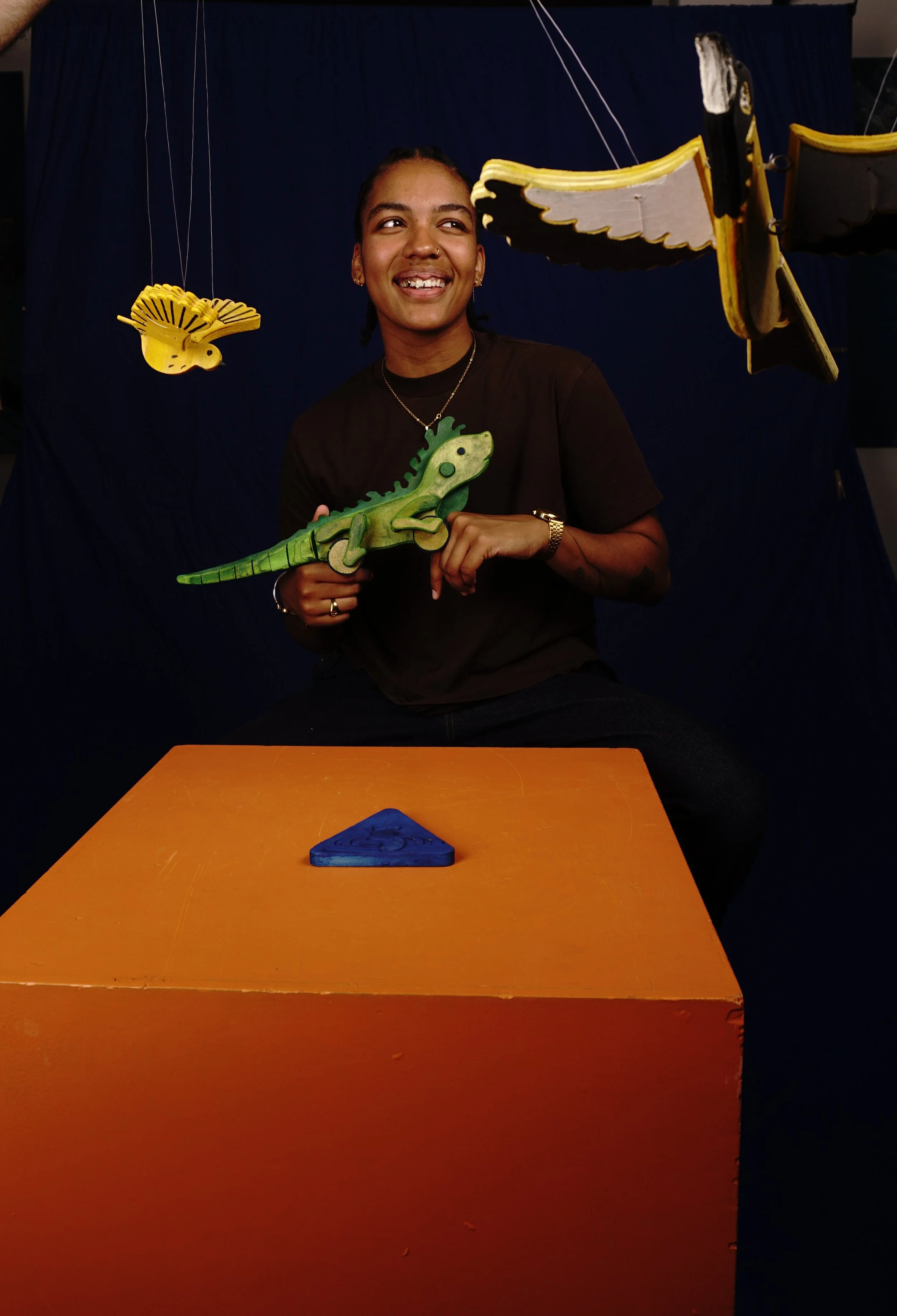 A woman smiling and holding a green wooden lizard puppet, seated at an orange table with a blue triangular object in front. Hanging yellow and black butterflies and a yellow bird are in the background, with a dark blue backdrop.