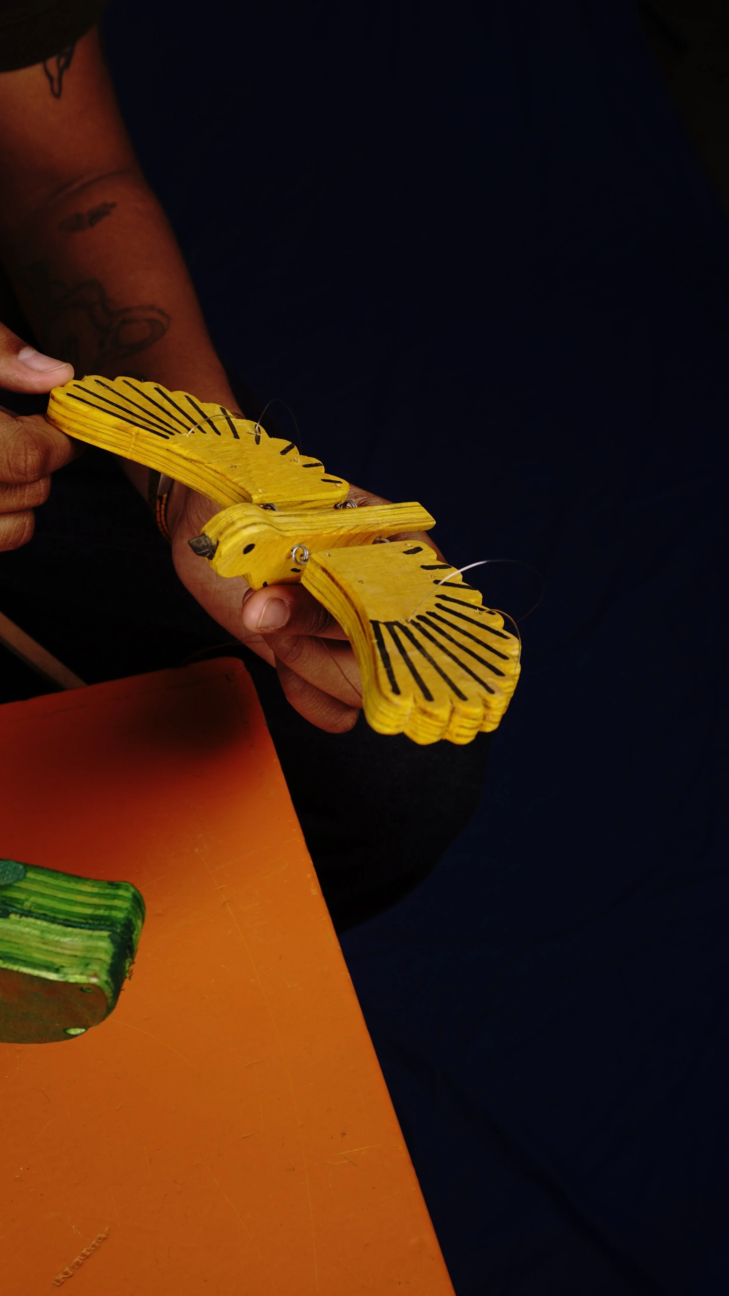 A person holding a yellow wooden butterfly craft with black lines, with part of a colorful book on an orange surface nearby.