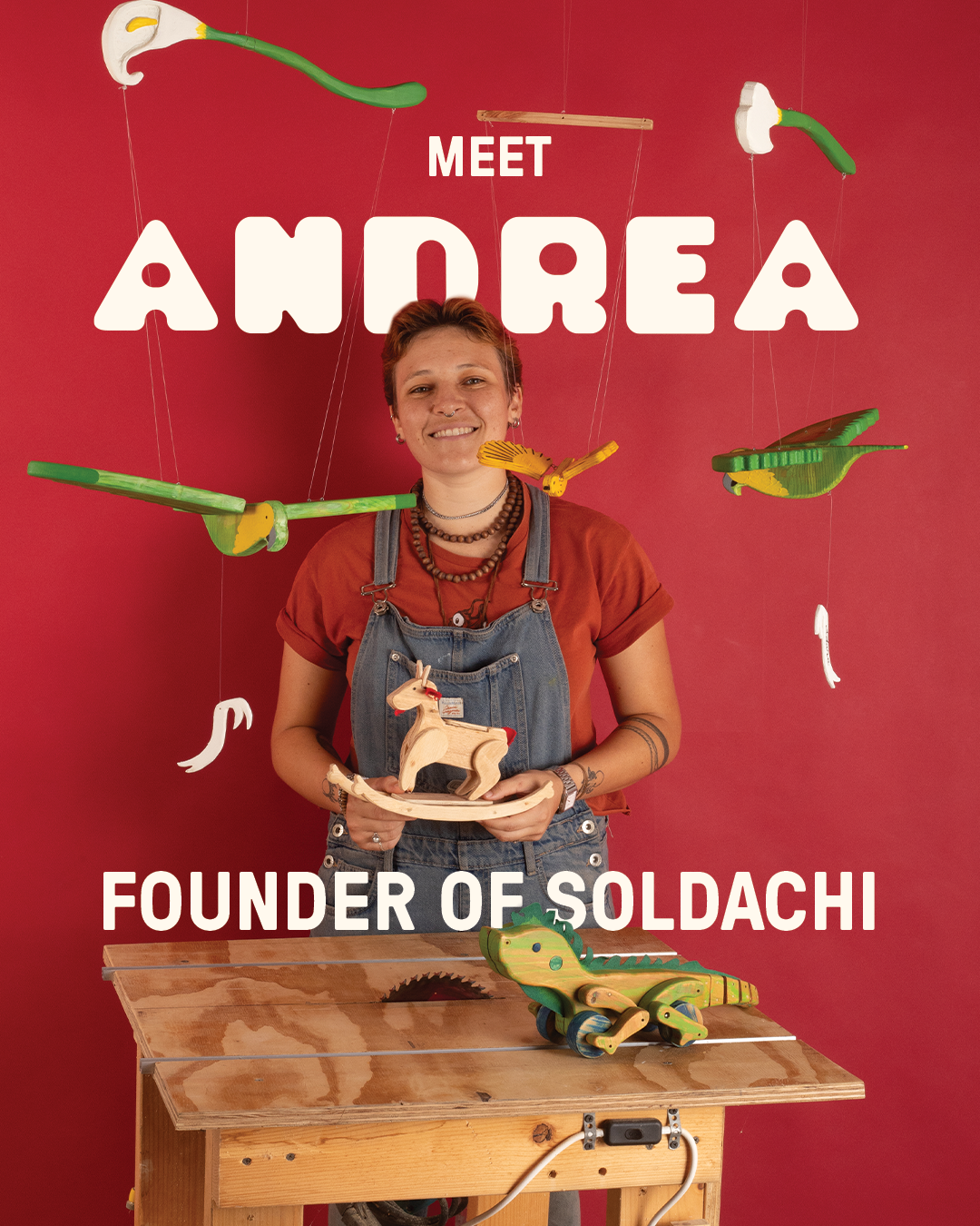 Portrait of Andrea, founder of Soldachi, smiling and holding a wooden rocking horse. She is standing in front of a red background with hanging colorful wooden toy animals and utensils, and there is a wooden table with a wooden crocodile toy in front of her.