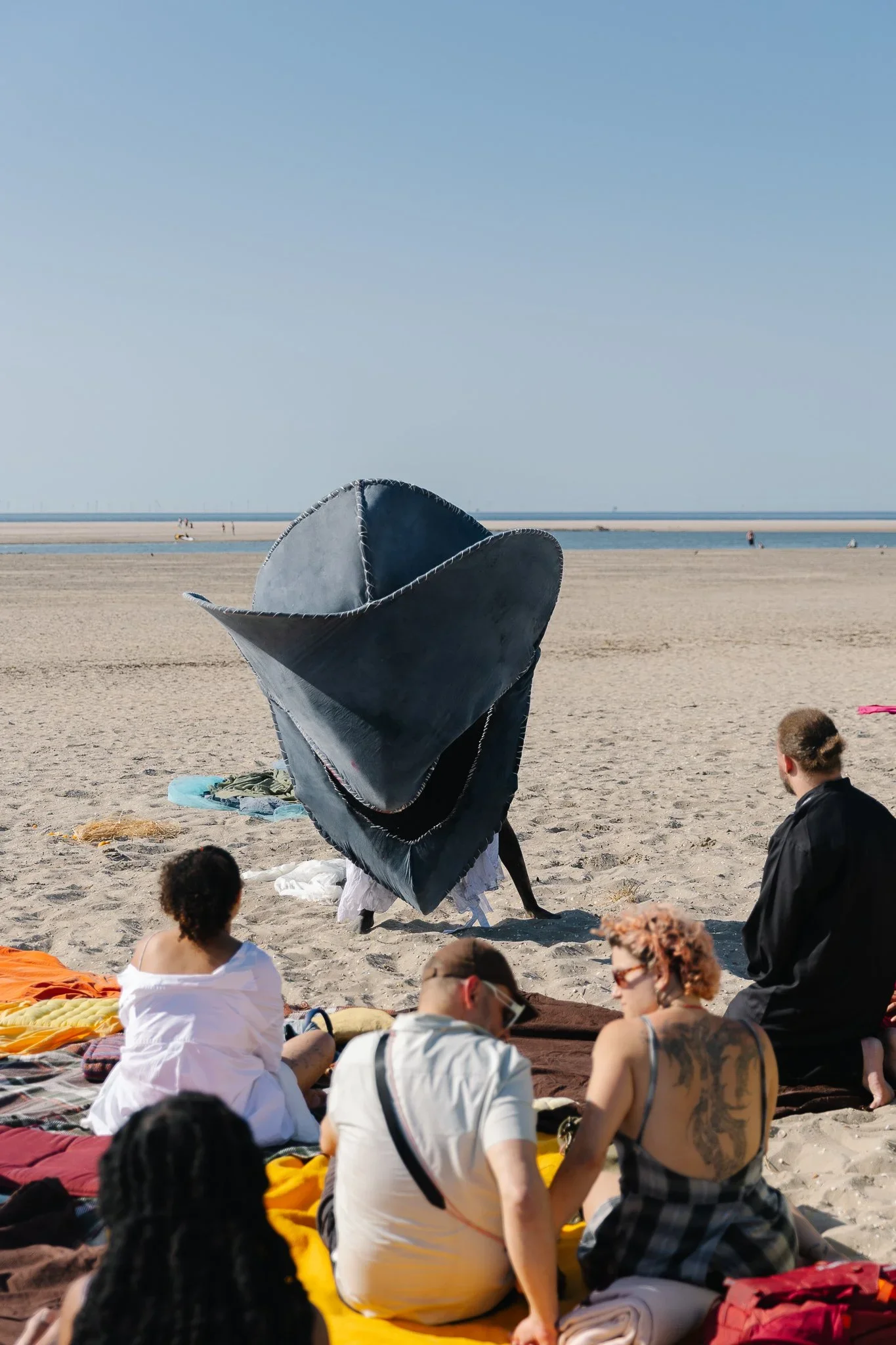 People sitting on the sand at the beach, with some standing and a large black hat in the center, and the ocean and sky in the background.