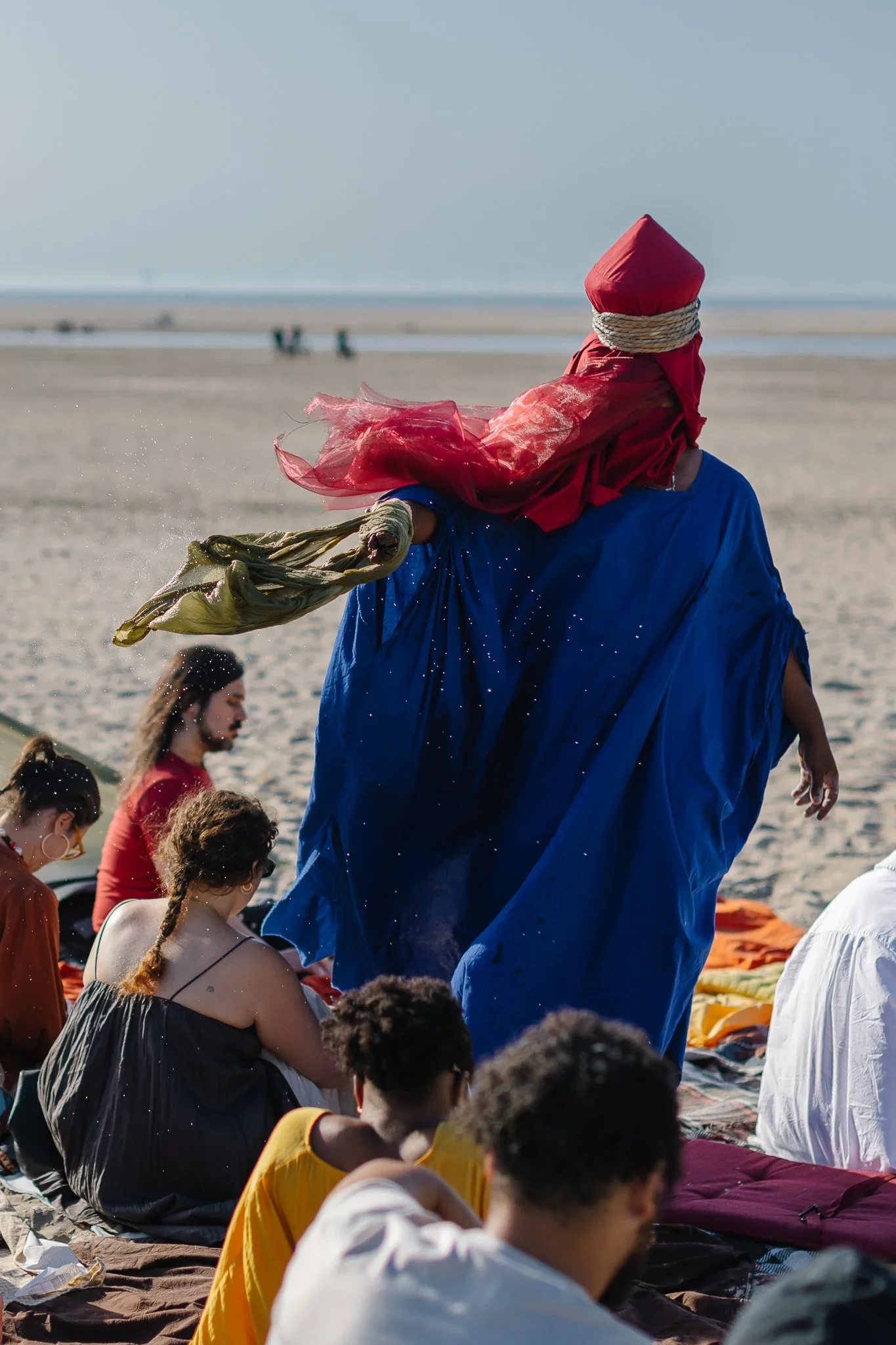 People sitting and meditating on a beach during sunrise or sunset, with a person in a blue robe and red hat standing in the background.