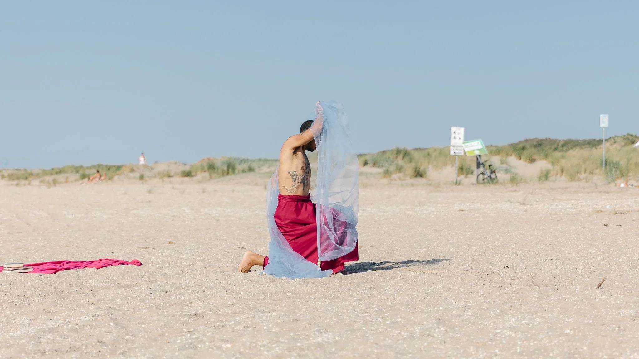 A person kneeling on a beach, holding a sheer fabric over their head with one hand. They are wearing red shorts and a tattoo is visible on their arm. There are a few items on the sand, including a pink towel or blanket, and signs and a bicycle can be seen in the background.