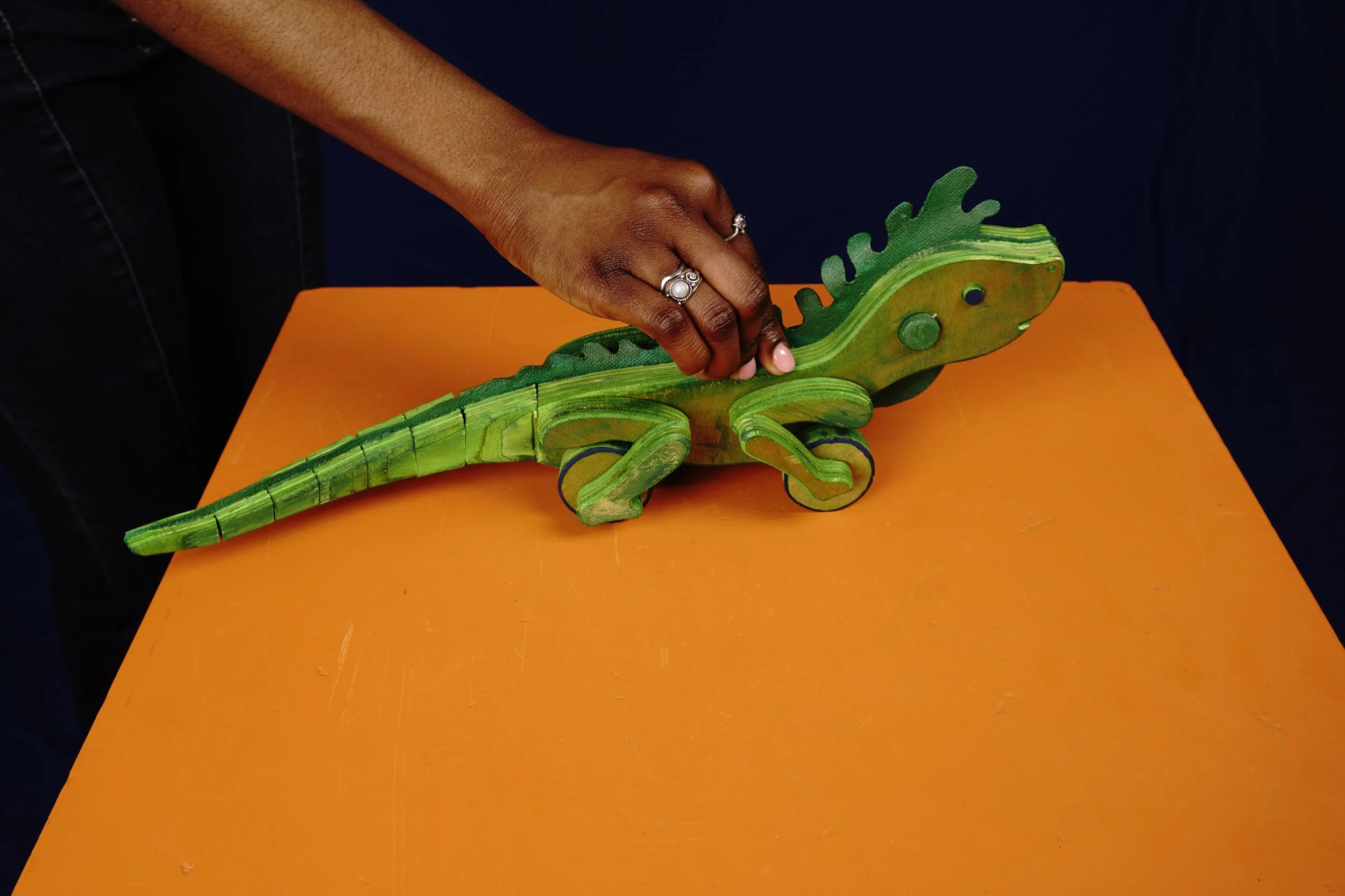 A person with dark skin is touching a colorful wooden chameleon toy on an orange table, wearing rings on their fingers. The background is dark.