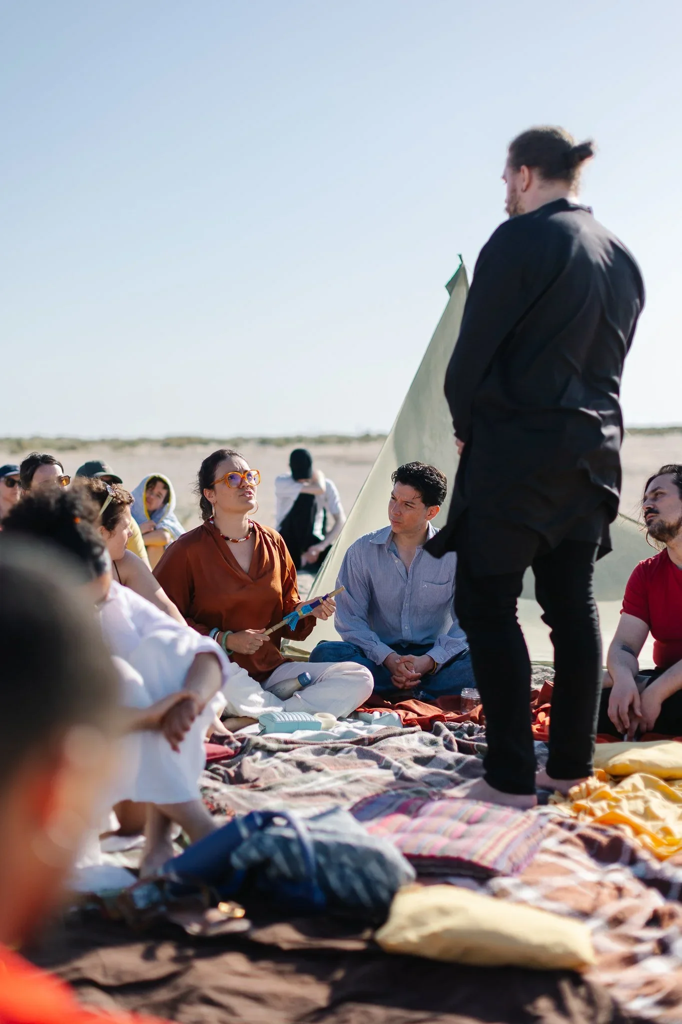 A group of people sitting on blankets outdoors, listening to a person standing and speaking to them during daytime.