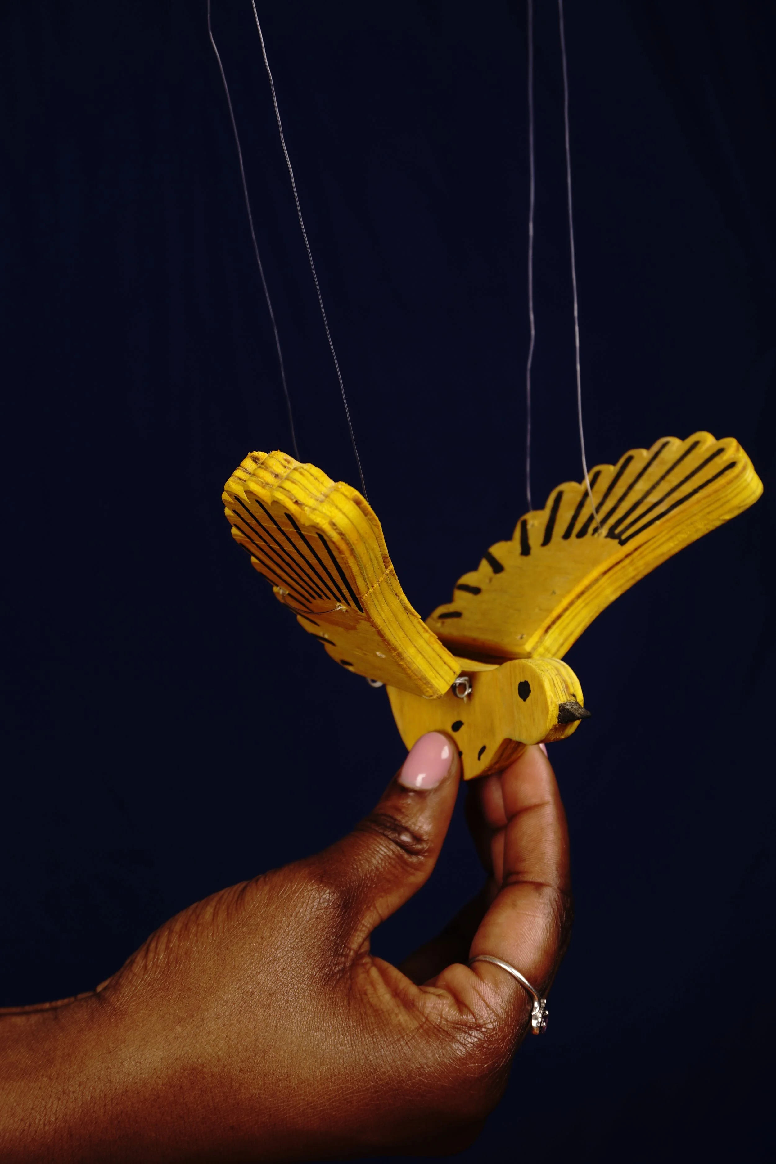 A person's hand holding a yellow wooden bird marionette with black painted details, suspended by thin strings against a dark background.