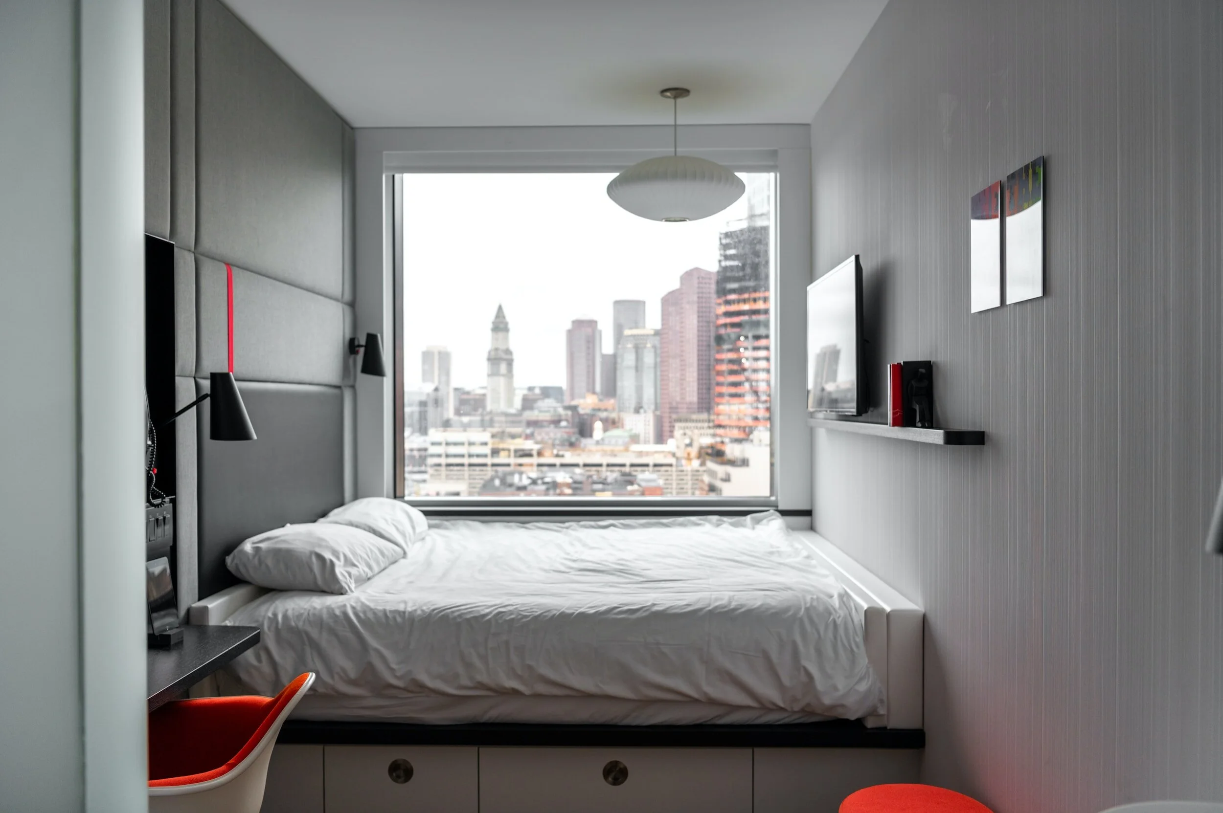 Modern hotel room with a large window showing a cityscape with tall buildings, a bed with white bedding, a mounted TV, and minimalistic decor.