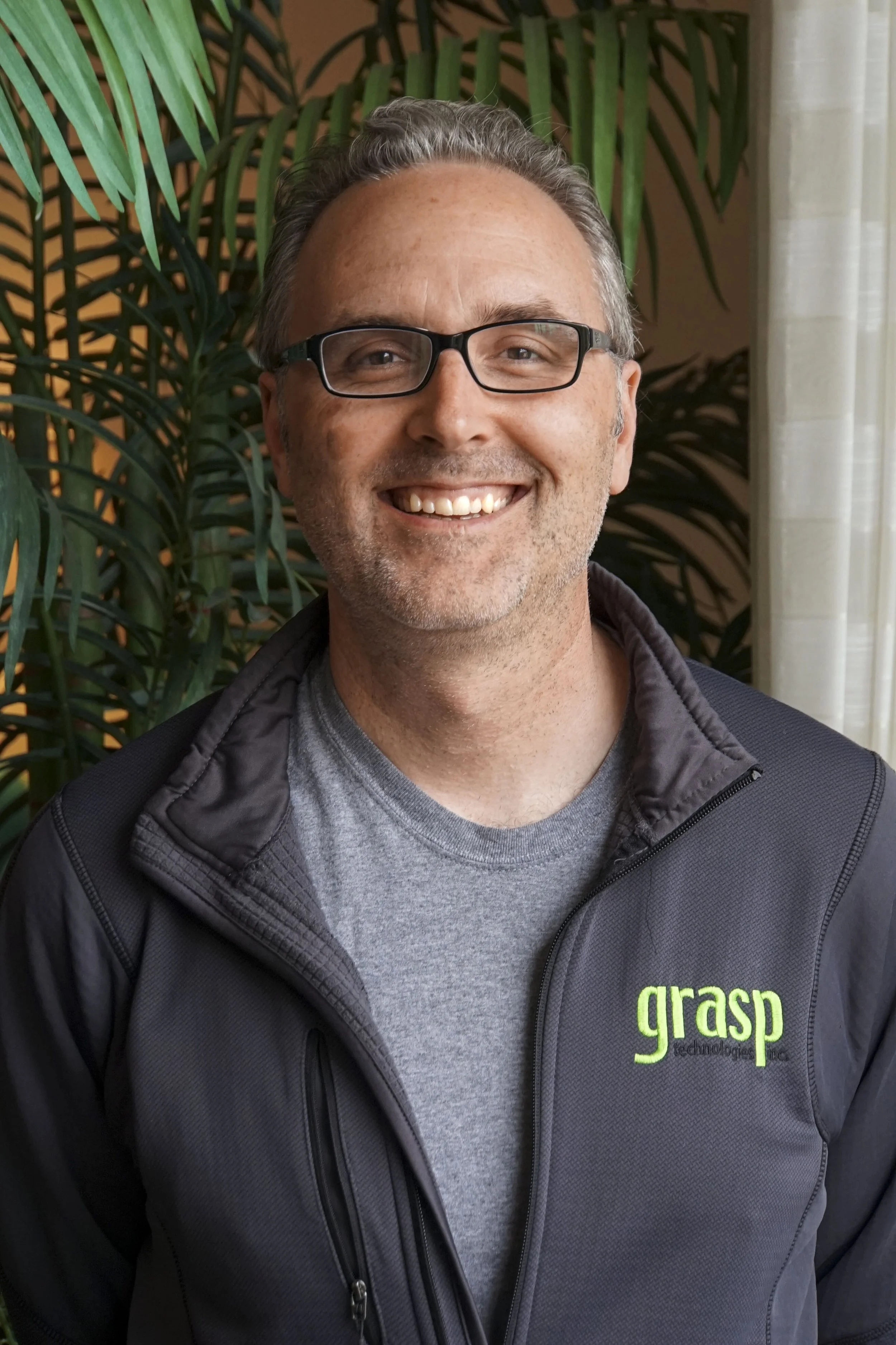 Black and white photo of a man wearing glasses, smiling, with a background of palm leaves.