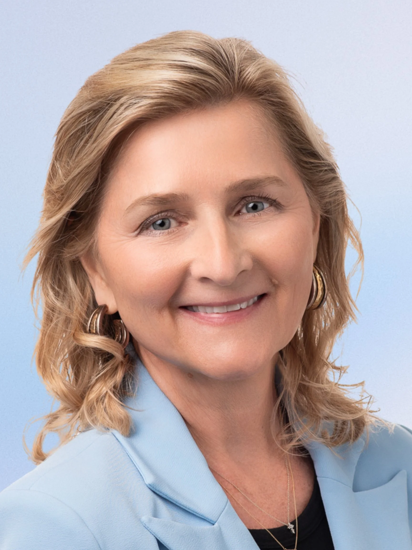 A black and white portrait of a woman with shoulder-length wavy hair, wearing a light-colored blazer over a dark top, smiling at the camera.