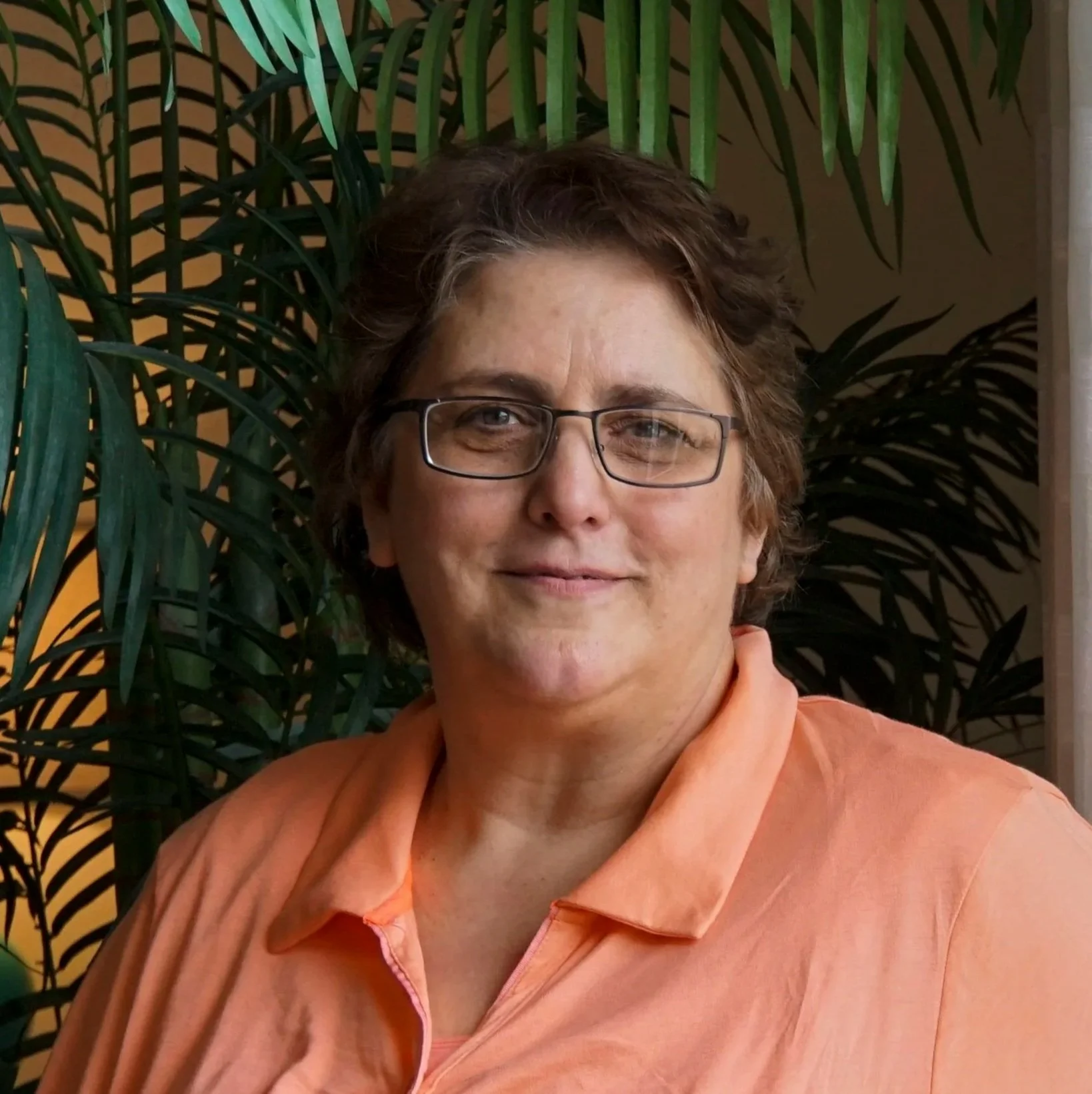 A woman with glasses and short curly hair smiling in front of leafy plants.