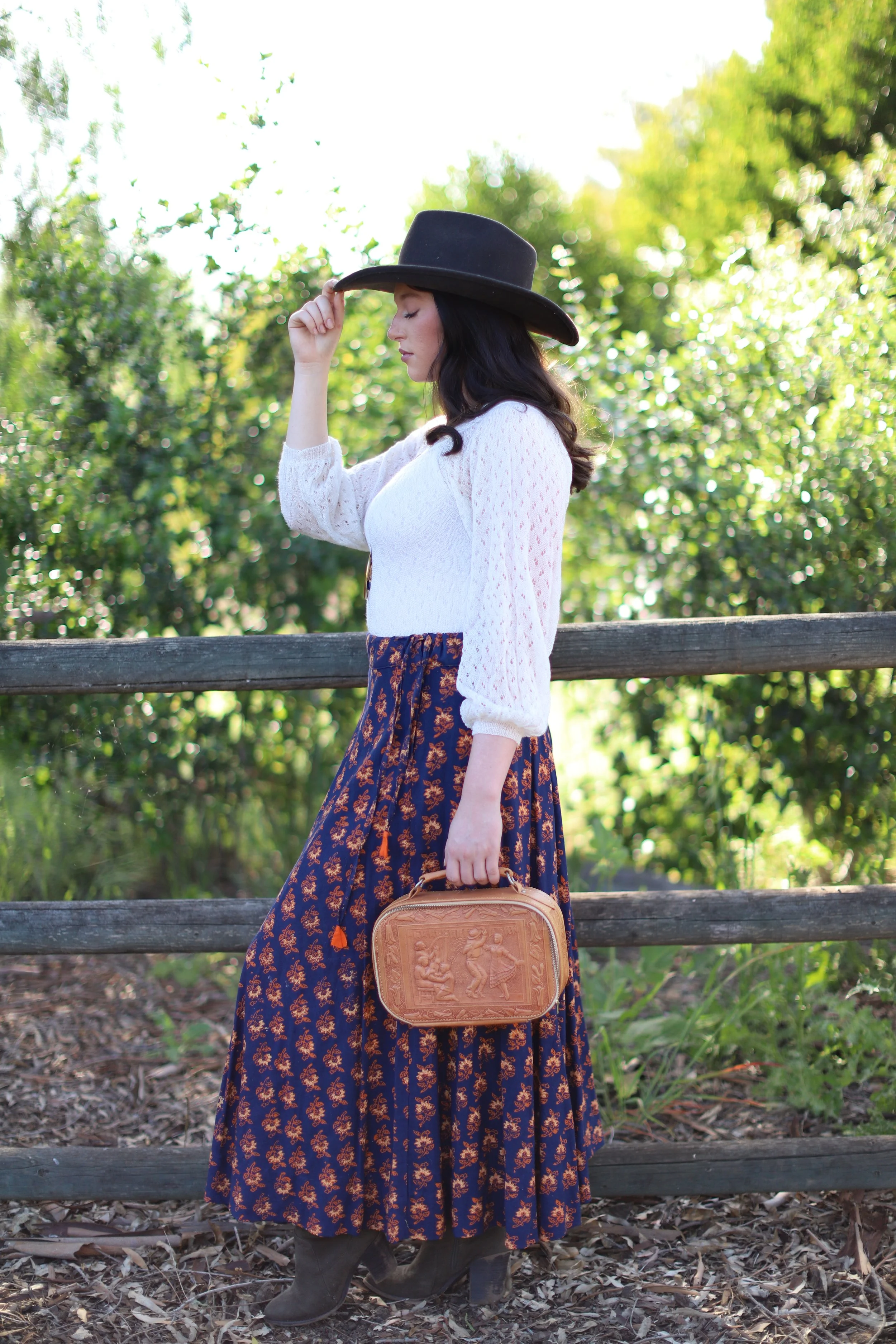 A woman in a black hat, white blouse, long patterned skirt, and brown boots stands outdoors near a wooden fence, holding a decorative handbag, with greenery and sunlight in the background.