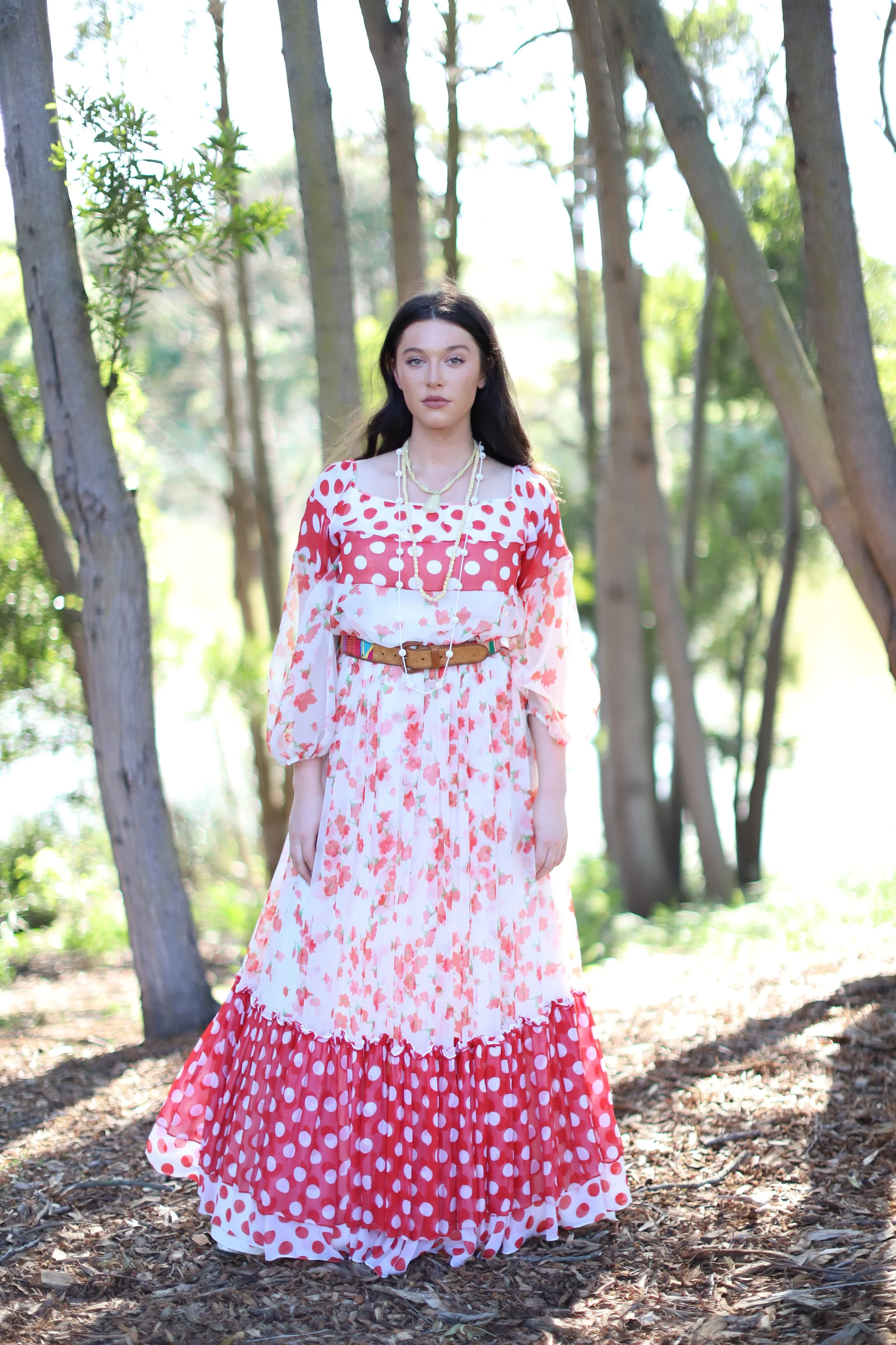 A woman standing outdoors among trees, wearing a long, flowing dress with red and pink floral patterns and polka dots, accessorized with layered necklaces and a brown waist belt.