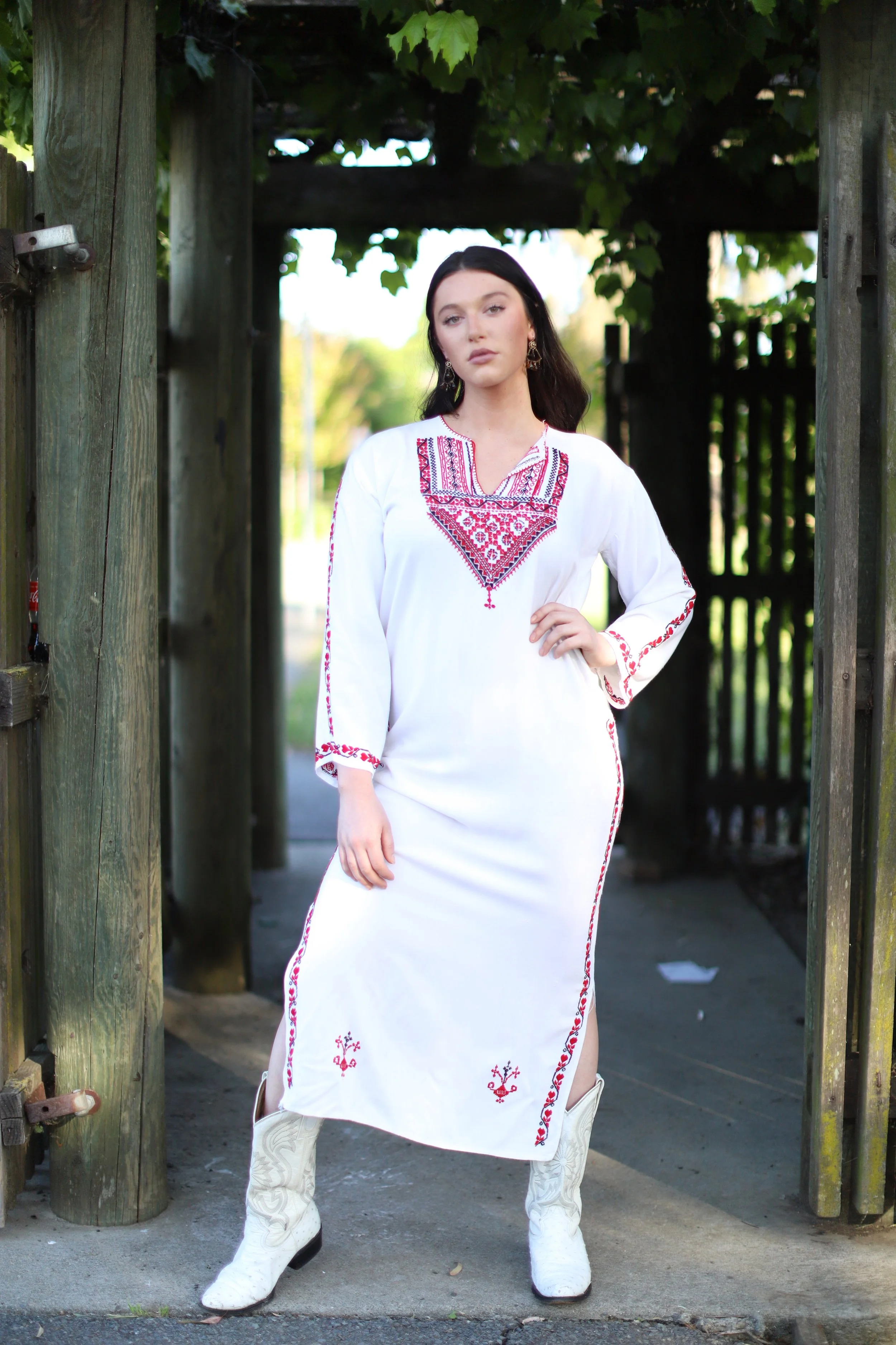 A woman wearing a white embroidered dress and cowboy boots standing under a wooden archway with greenery overhead.