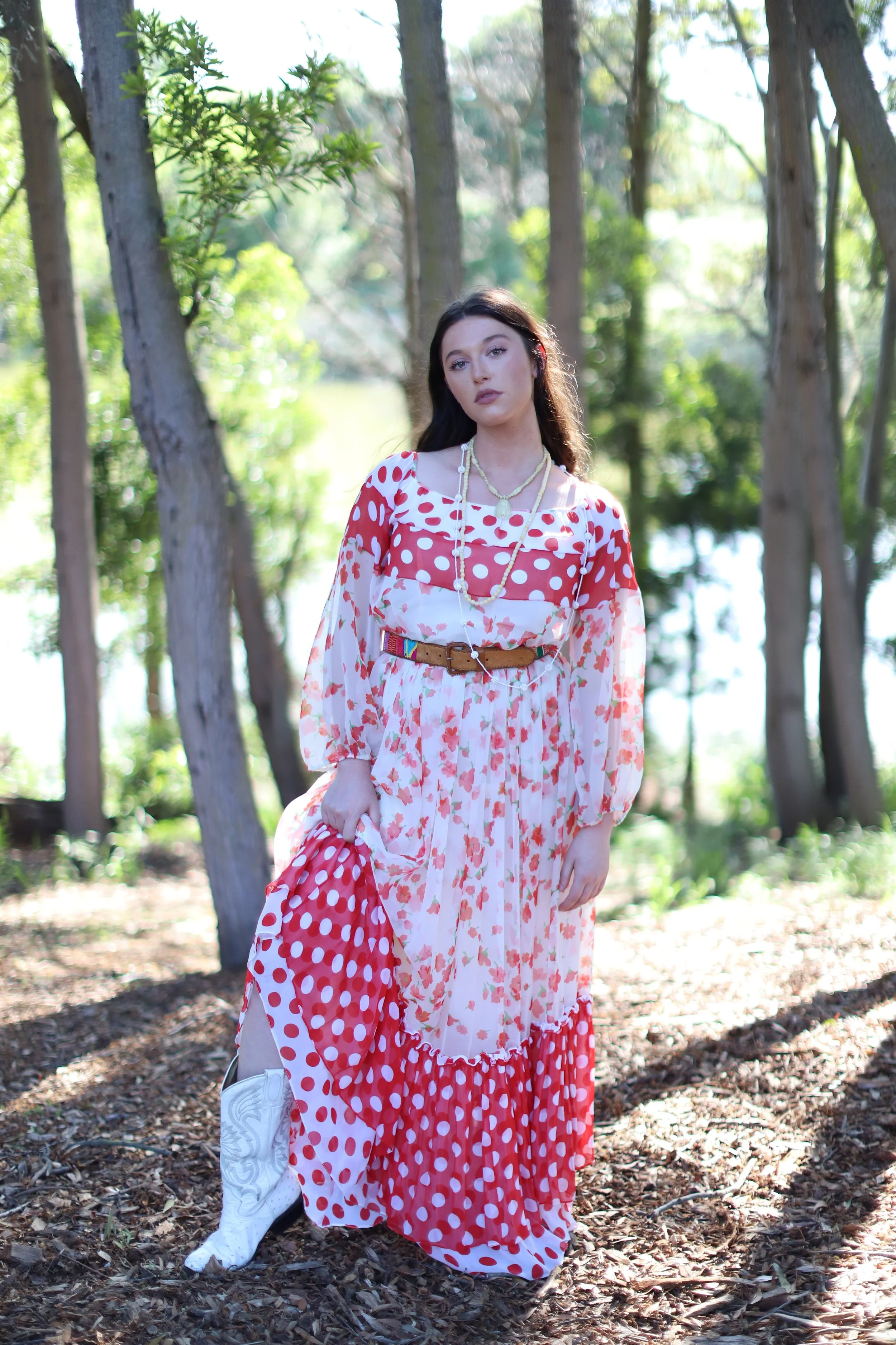 A woman with long dark hair in a floral and polka dot red and white dress standing in a wooded area with trees and sunlight filtering through.