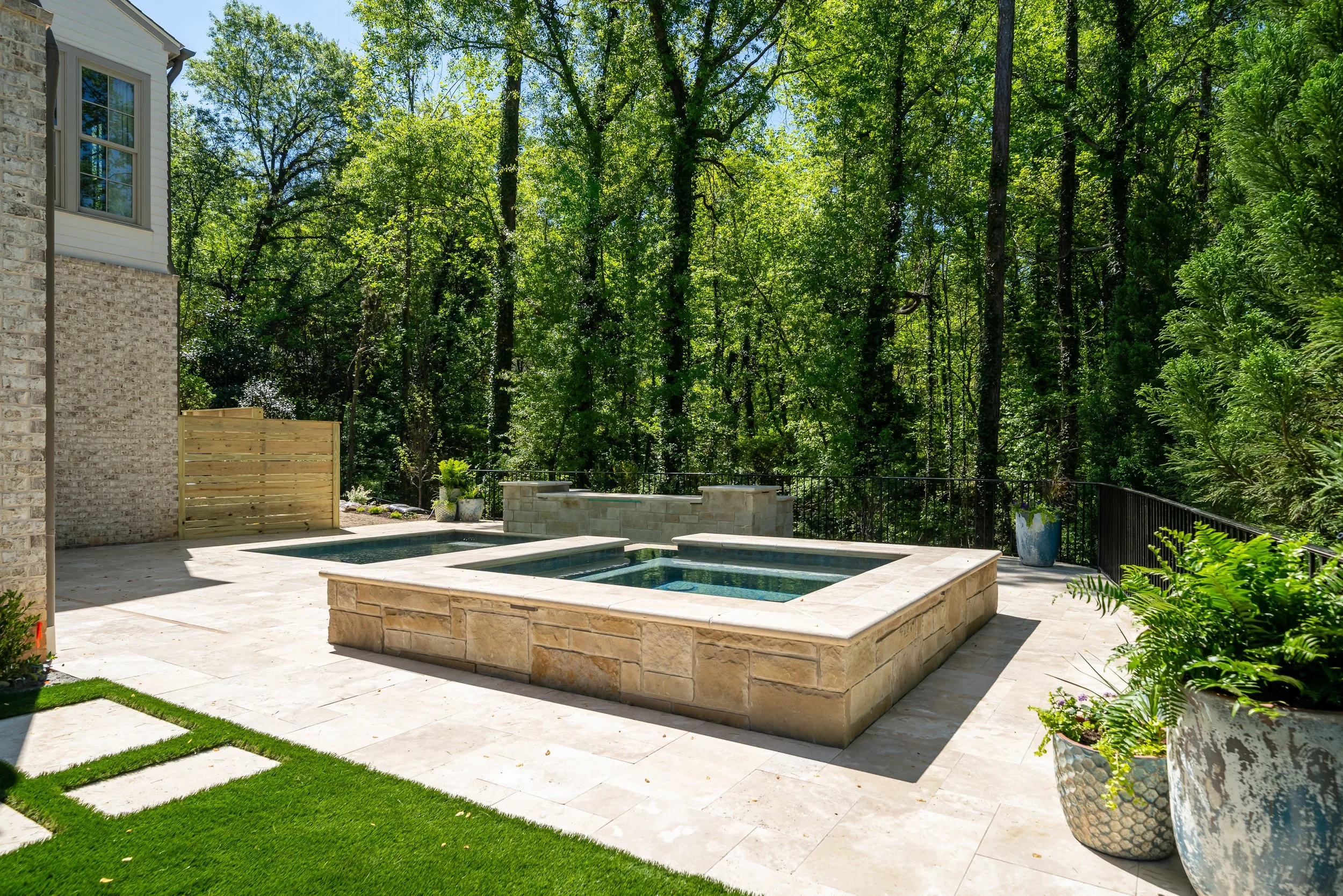 Backyard patio with a hot tub and pool, surrounded by potted plants and a fence, backed by a wooded area with tall trees.