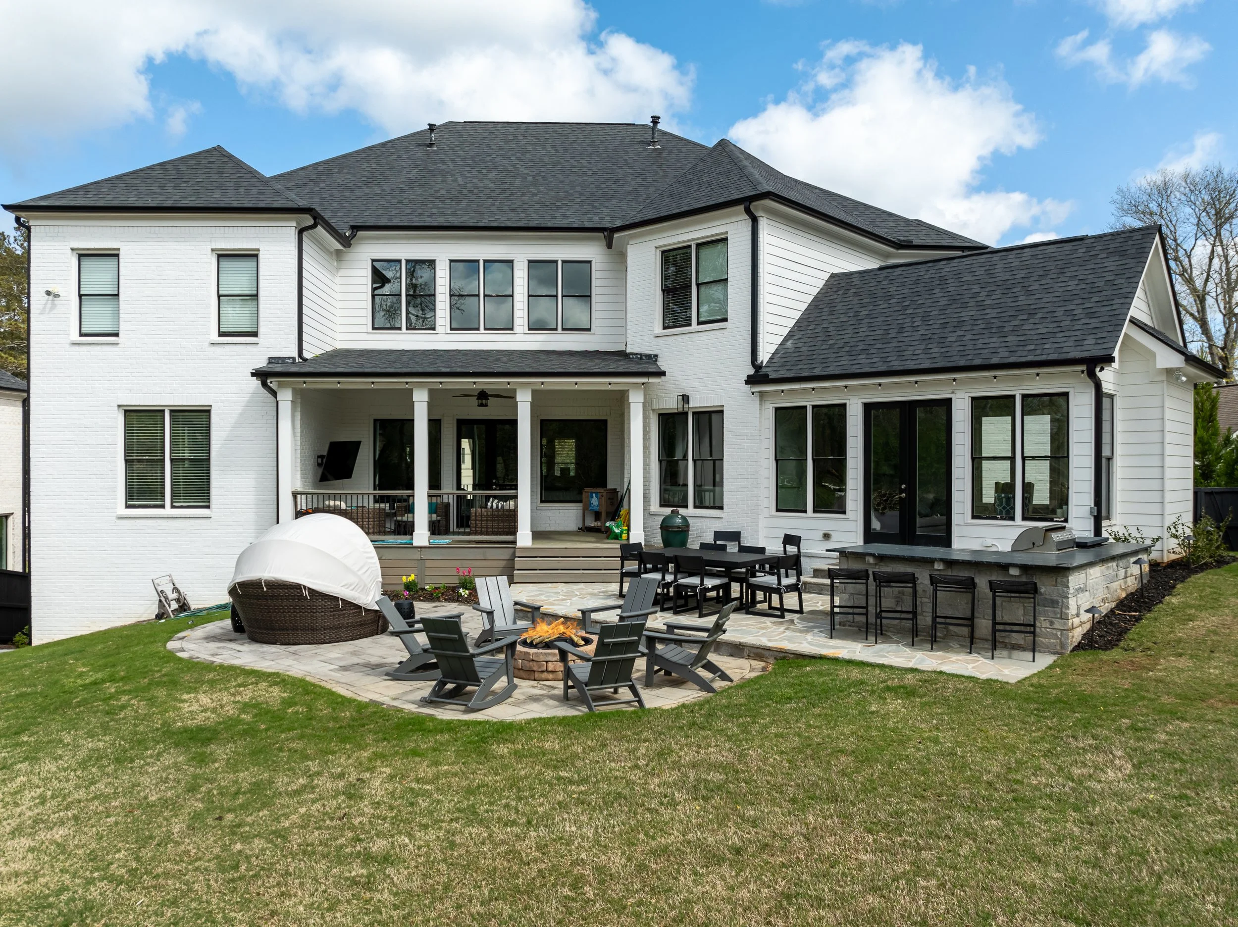 Modern white two-story house with black roof, large back porch, outdoor seating area, fire pit, and yard with grass and trees.