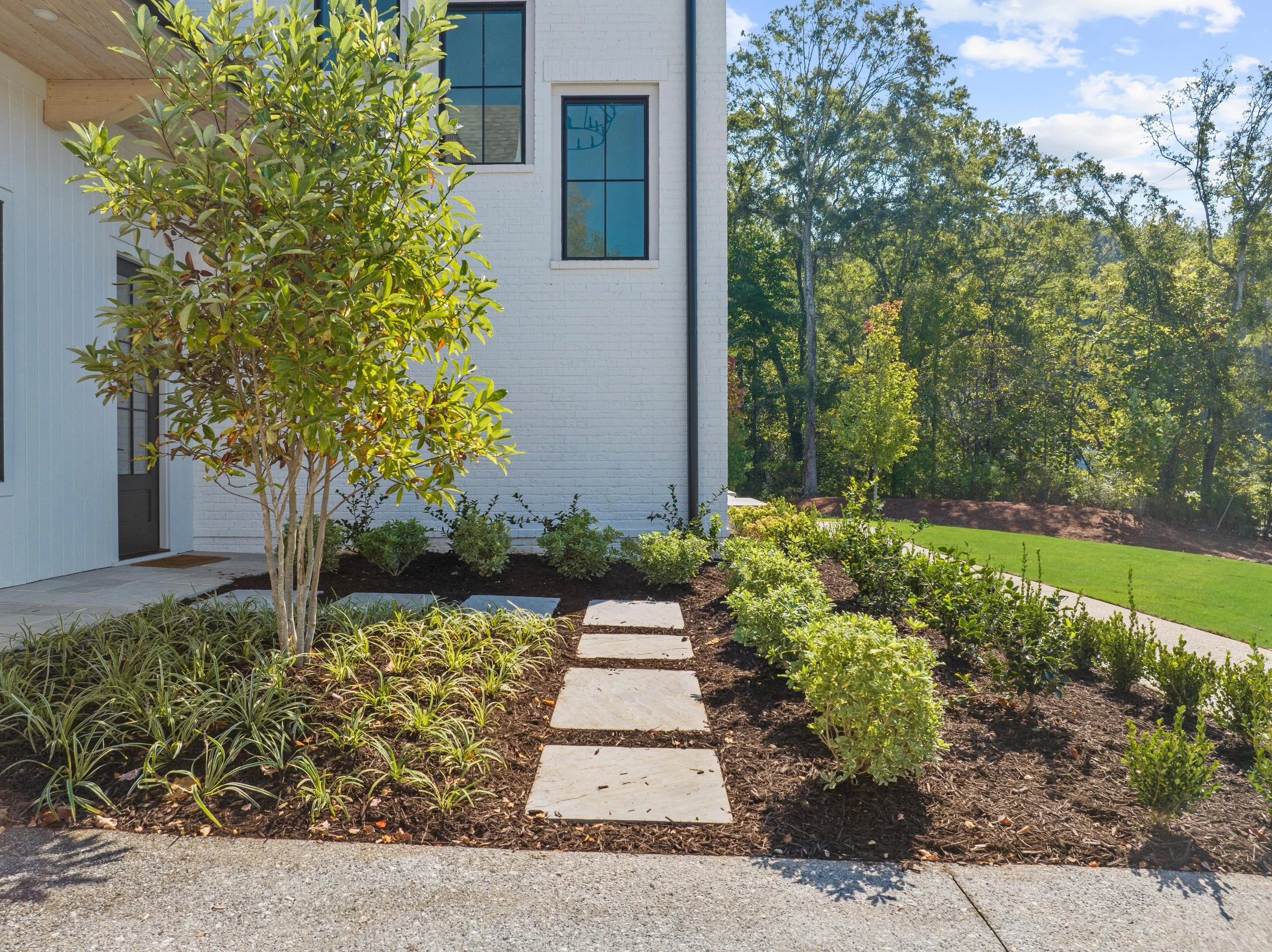 Side yard with a white brick house, concrete stepping stones, landscaped garden with various shrubs and plants, mature tree, green lawn, trees in the background, and a partly cloudy sky.