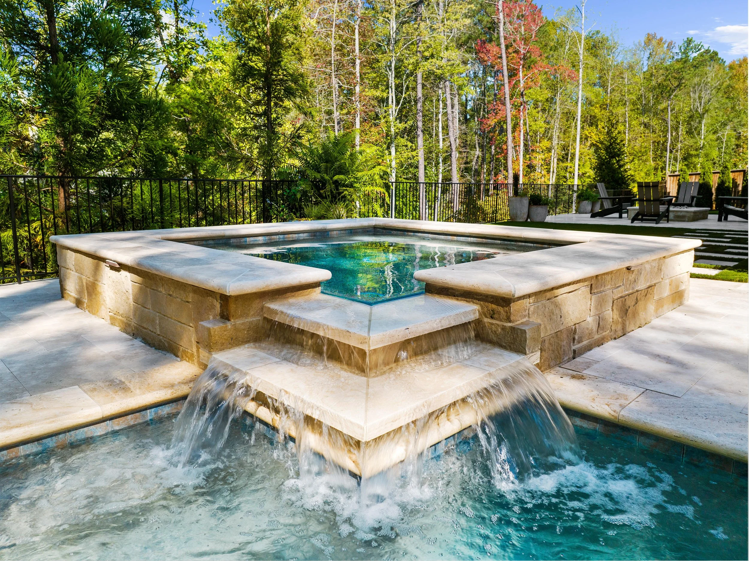 A stone hot tub with water cascading over the edge, situated outdoors on a patio surrounded by trees and chairs.