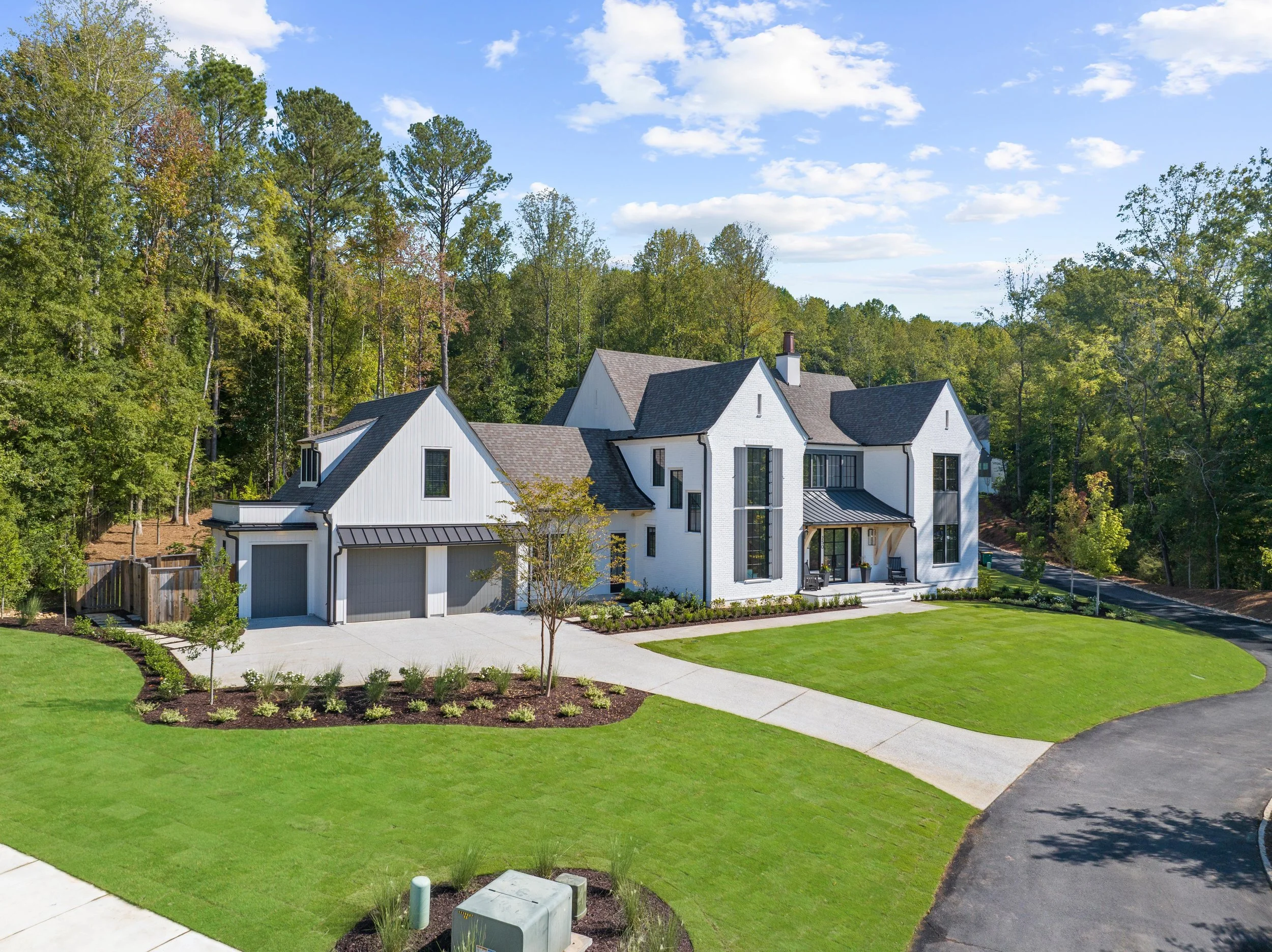 Modern white house with multiple gabled roofs, large windows, and a front porch, surrounded by green lawn and trees, under a partly cloudy sky.