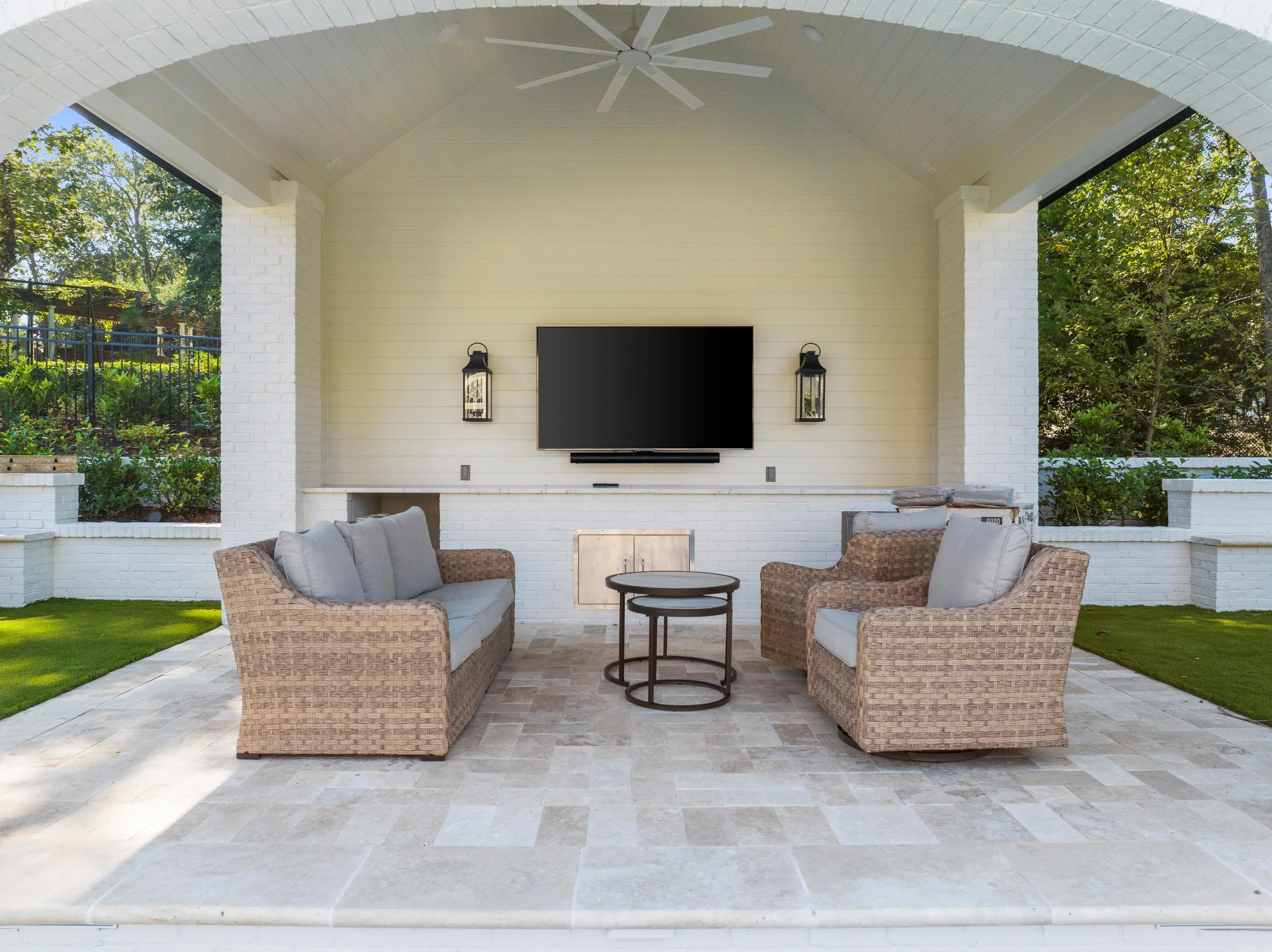 Outdoor living space with wicker sofa and armchair, round tables, wall-mounted TV, two wall lanterns, and white brick structure, surrounded by greenery.