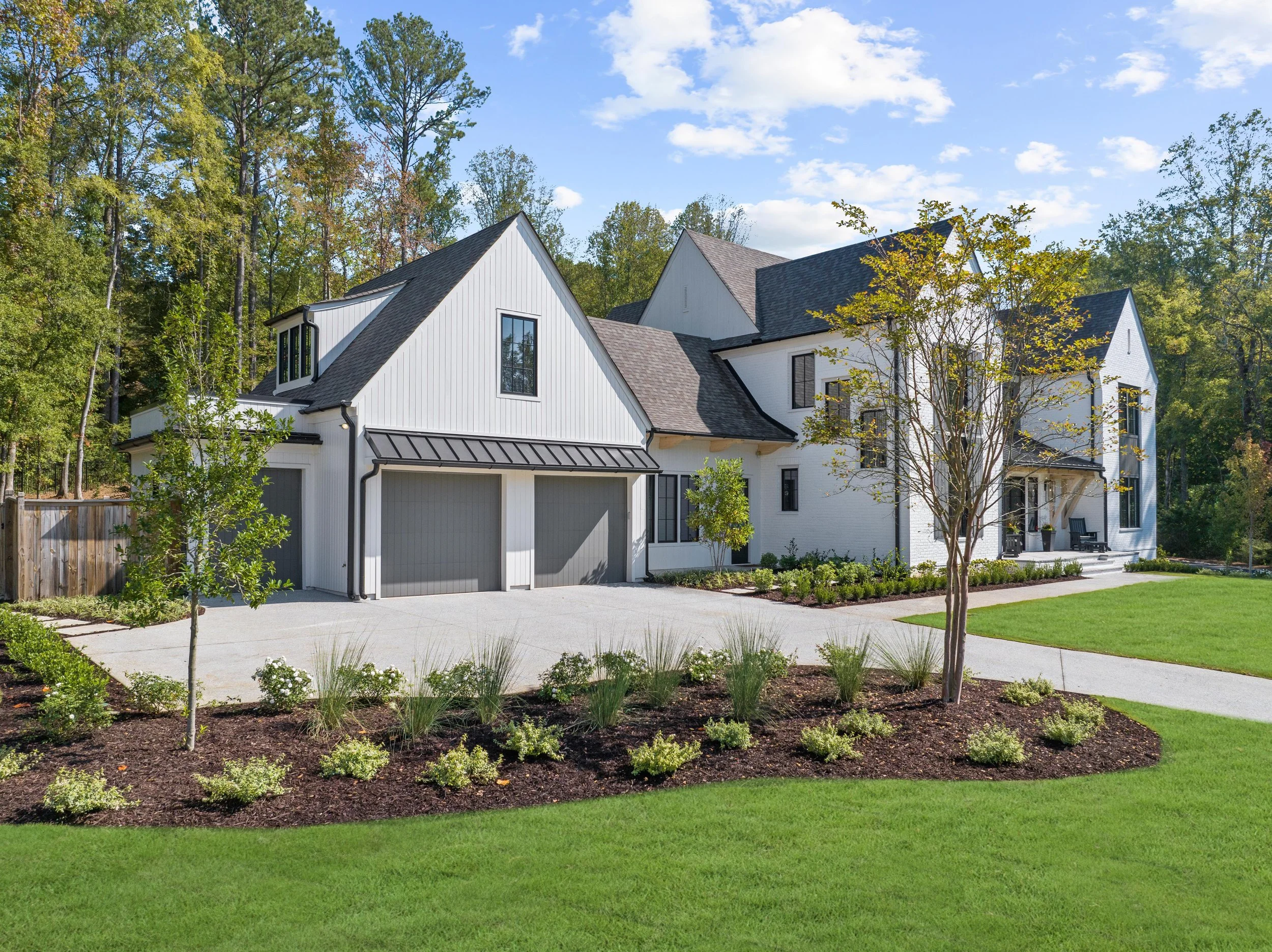 Modern white house with black accents, attached garage, surrounded by landscaped yard with trees, shrubs, and well-kept lawn, against a backdrop of trees and blue sky.