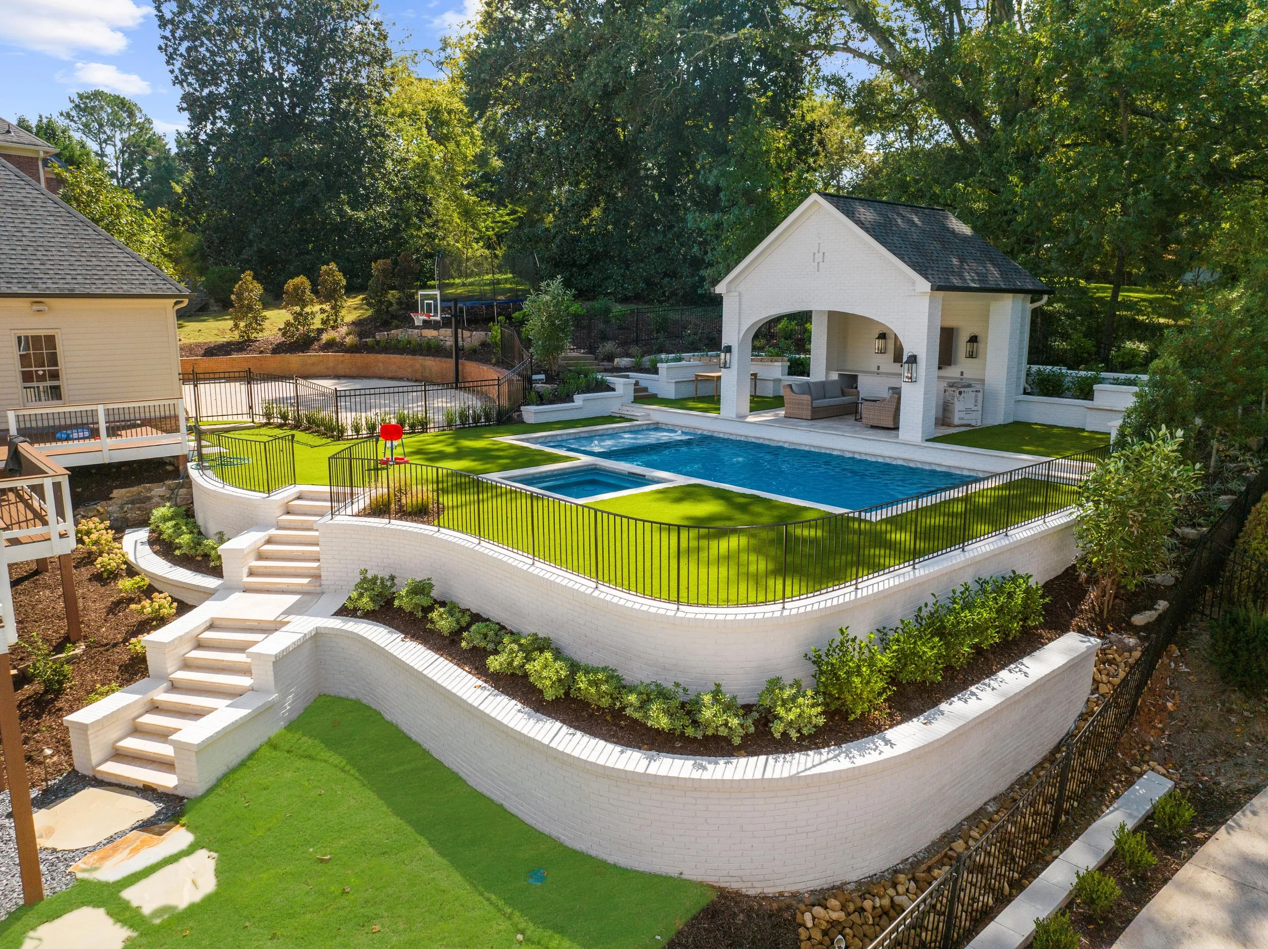 A backyard featuring a multi-level white brick patio with a swimming pool, an outdoor seating area under a covered pavilion, a basketball hoop, and a tennis court surrounded by lush green trees.