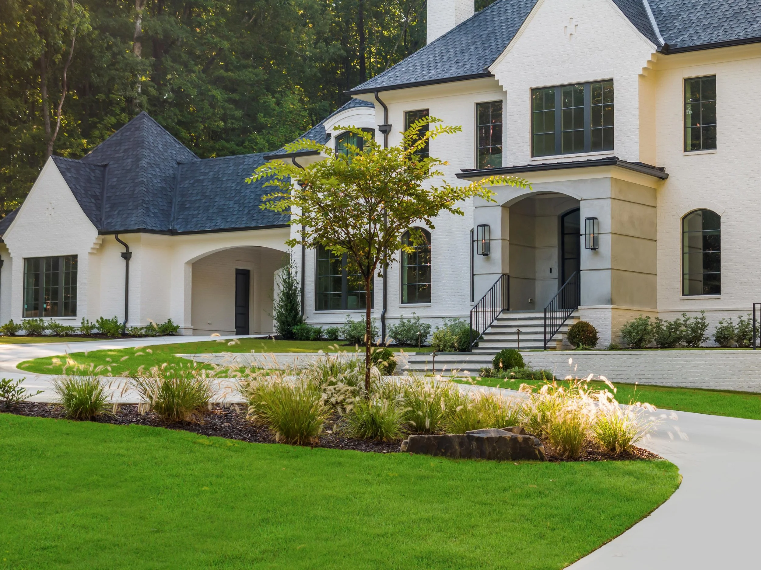 Large white house with black roof, front steps, and black railing, surrounded by landscaped yard with green grass, plants, and trees.