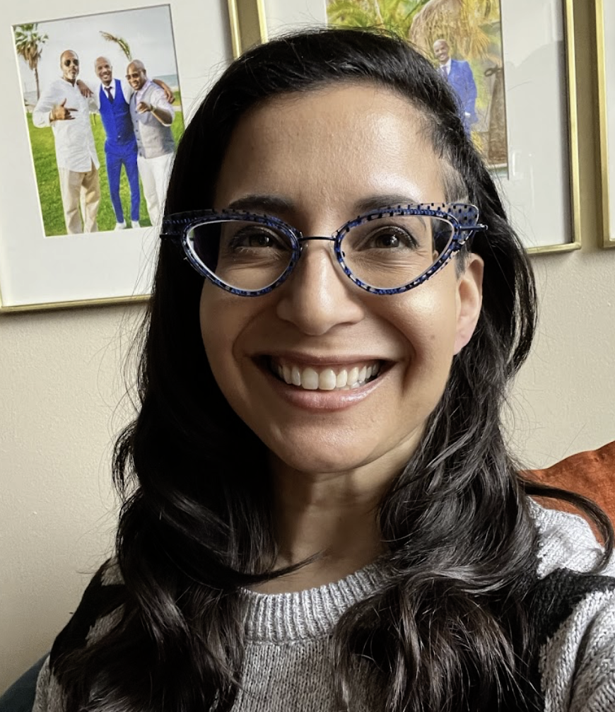 A woman with long dark hair and glasses with blue patterned frames smiling in front of framed family photos on a wall.