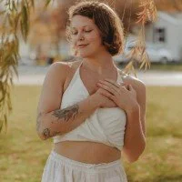 A young woman with short curly hair and tattoos, wearing a white sleeveless top and light-colored pants, standing outdoors with her eyes closed and her hands on her chest, in a serene setting with trees and a house in the background.