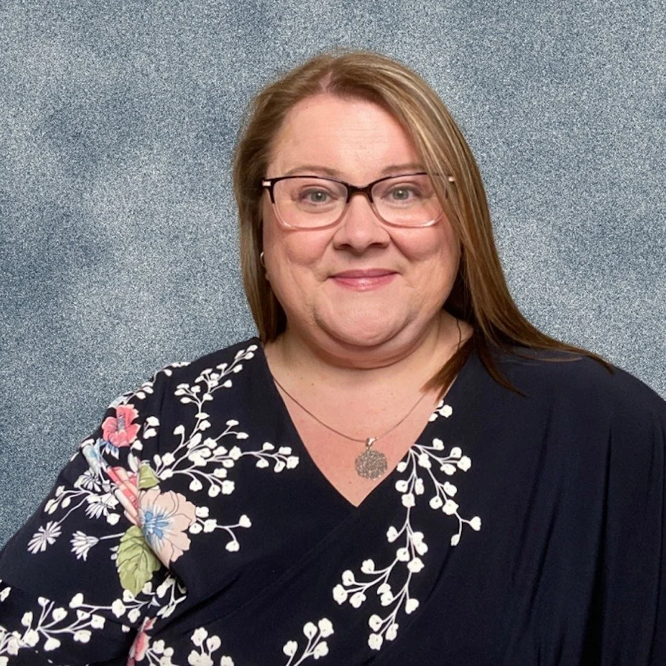 A woman with shoulder-length reddish-brown hair, wearing glasses, a black floral blouse, and a circular pendant necklace, standing in front of a textured gray background.