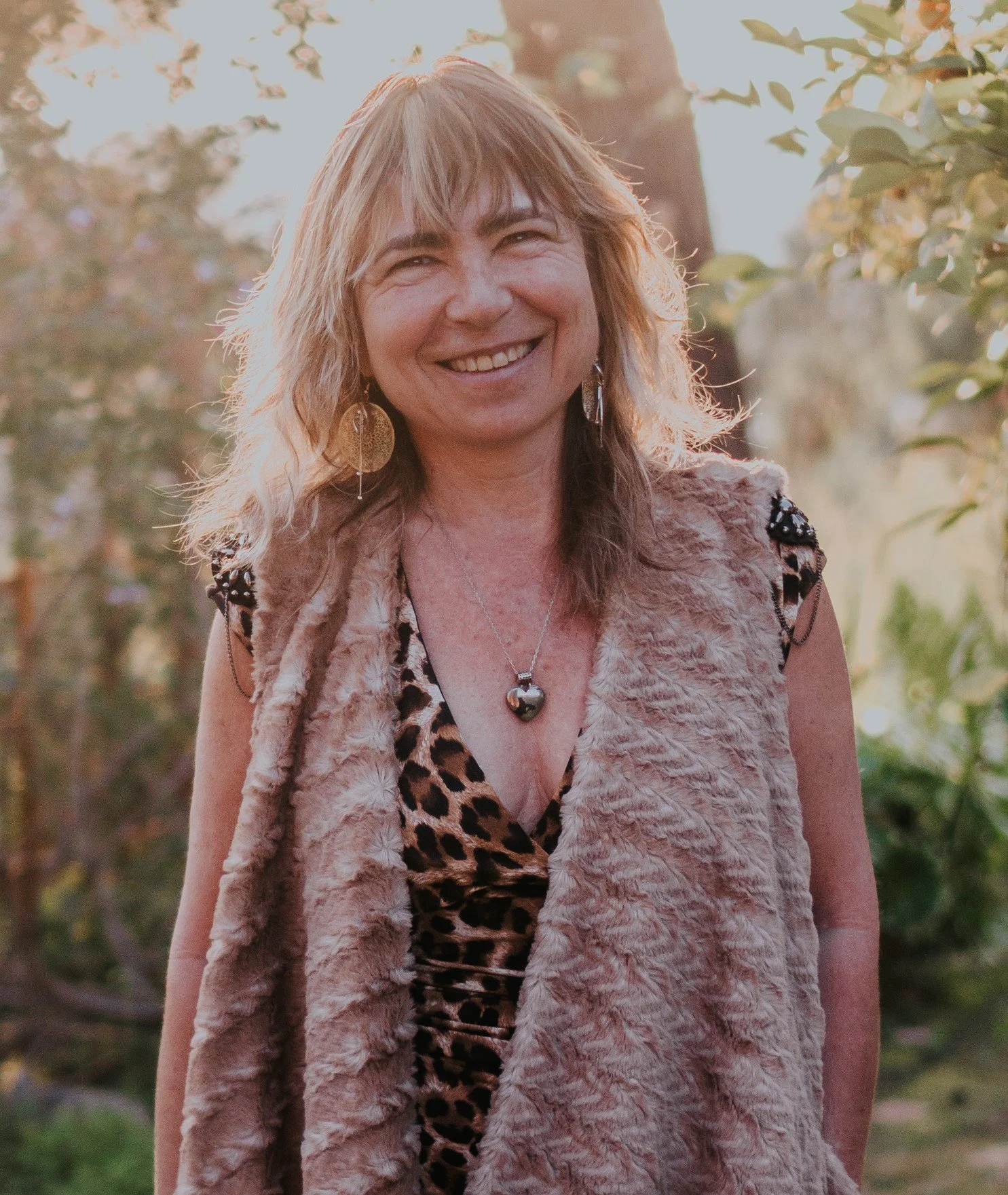 A smiling woman with wavy blonde hair stands outdoors in sunlight, wearing a leopard print top, a beige faux fur vest, and gold earrings.