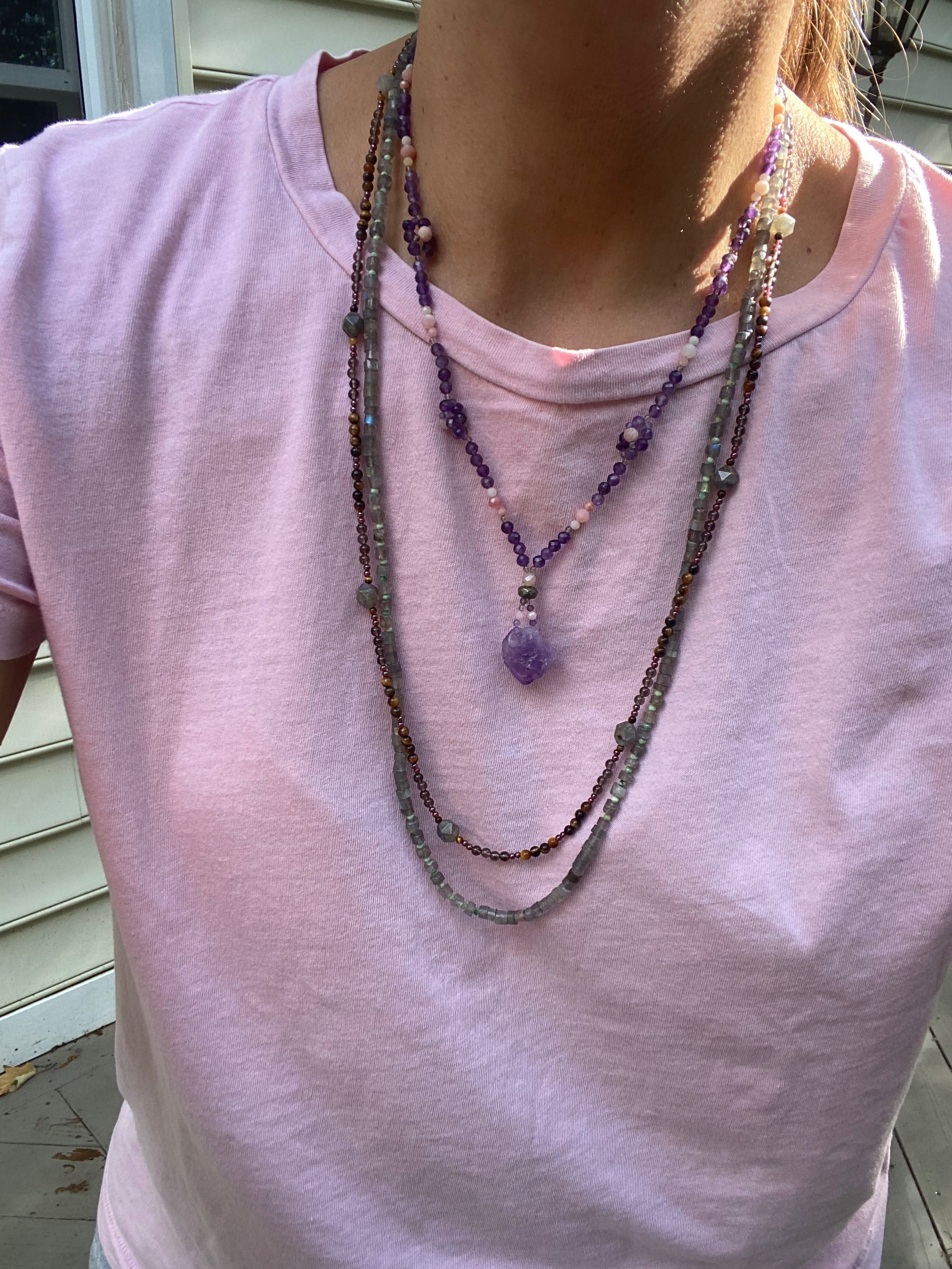 A raw faceted amethyst stone hangs on an 18 inch necklace of Amethyst and Pink Opal flowers. Layered with other long gemstone necklaces on a bright pink shirt