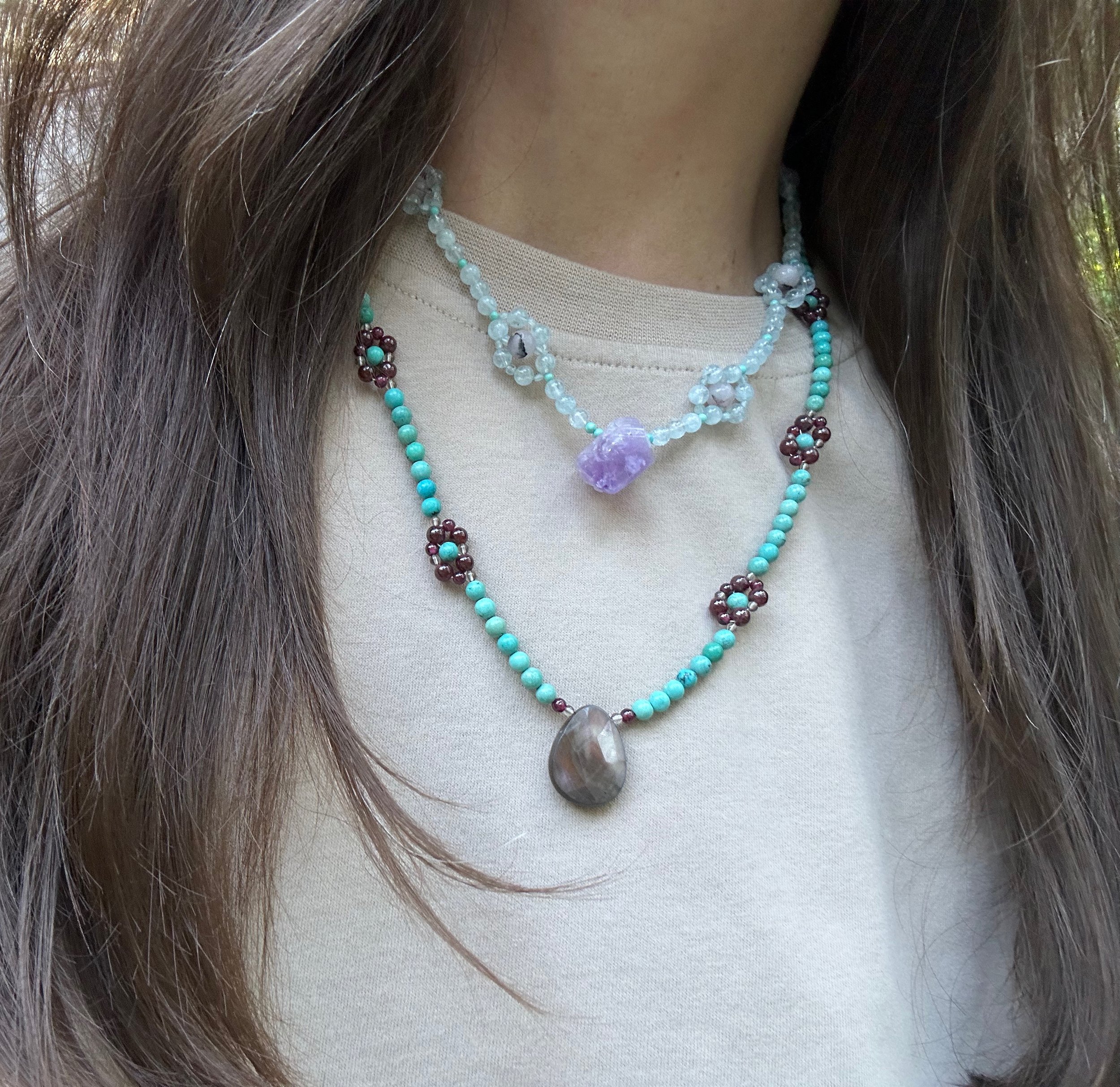 raw faceted amethyst stone dangles from a unique gemstone necklace of aquamarine and turquoise wildflowers and sits just below the collar bone on a white t-shirt. It is layered with a similar necklace