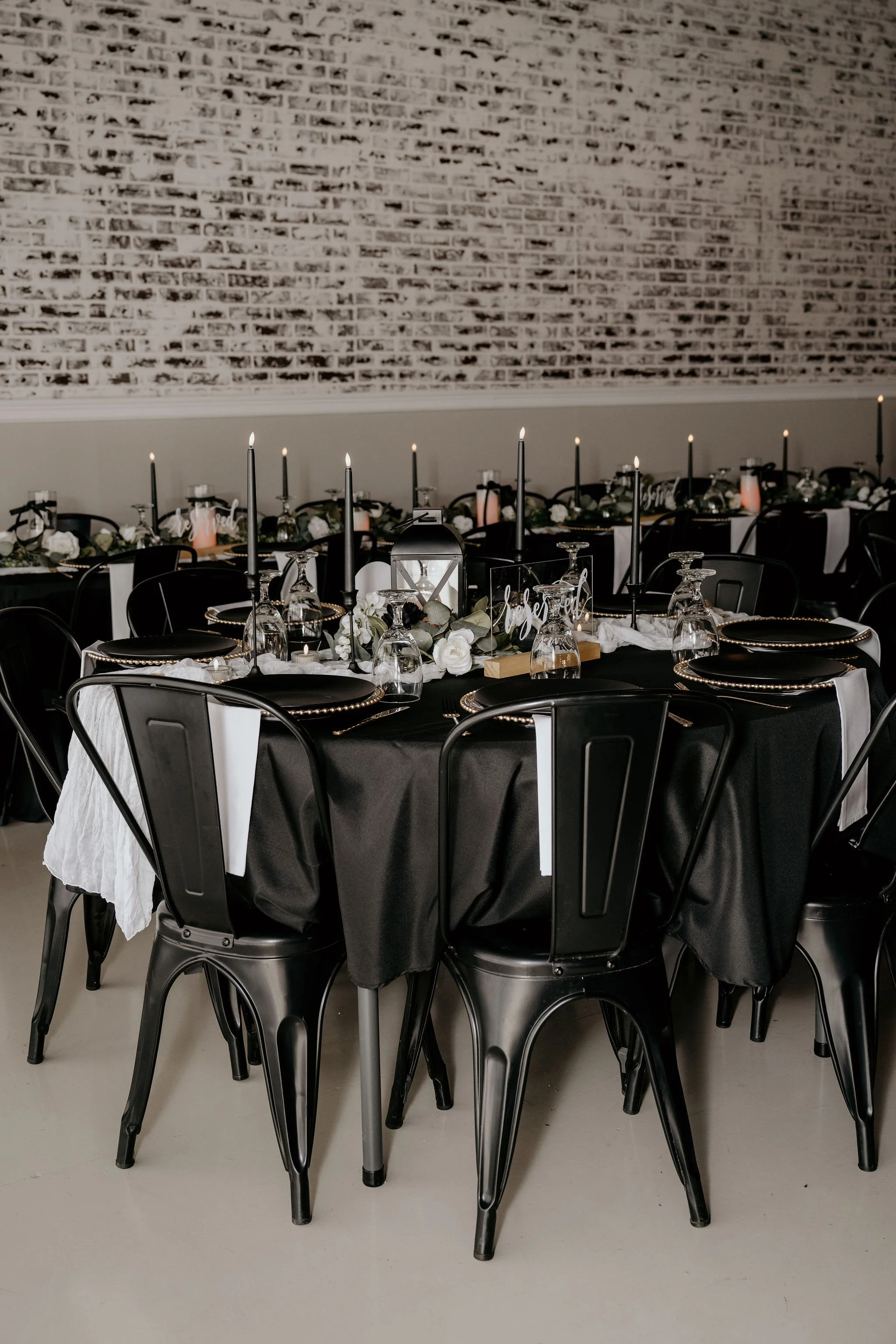 Elegantly set banquet table with black tablecloth, black metal chairs, white napkins, candles, wine glasses, and floral centerpieces in a room with a white brick wall.