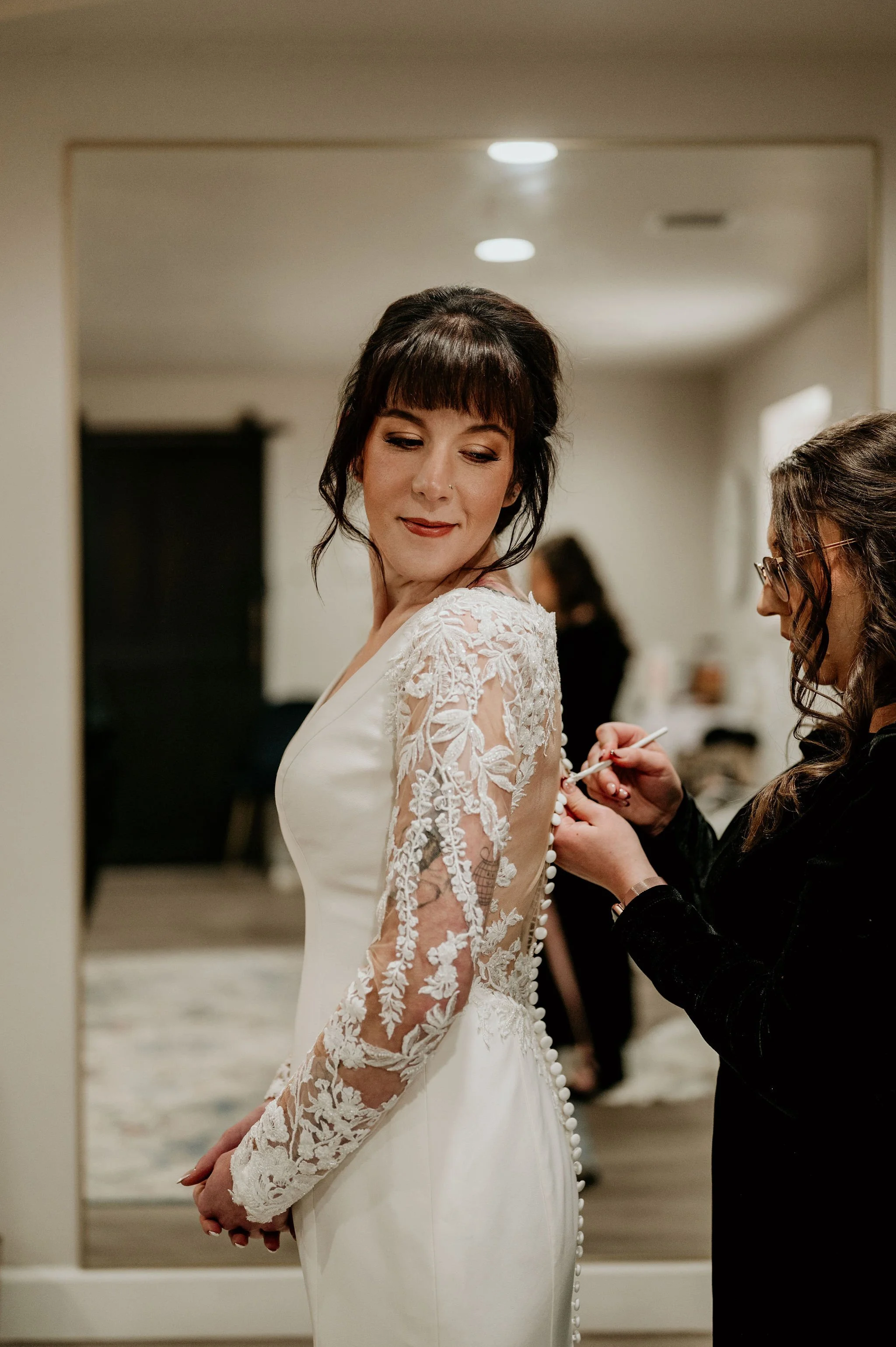 A bride in a white wedding dress with lace sleeves is getting ready, while a woman helps button up the back of her dress.