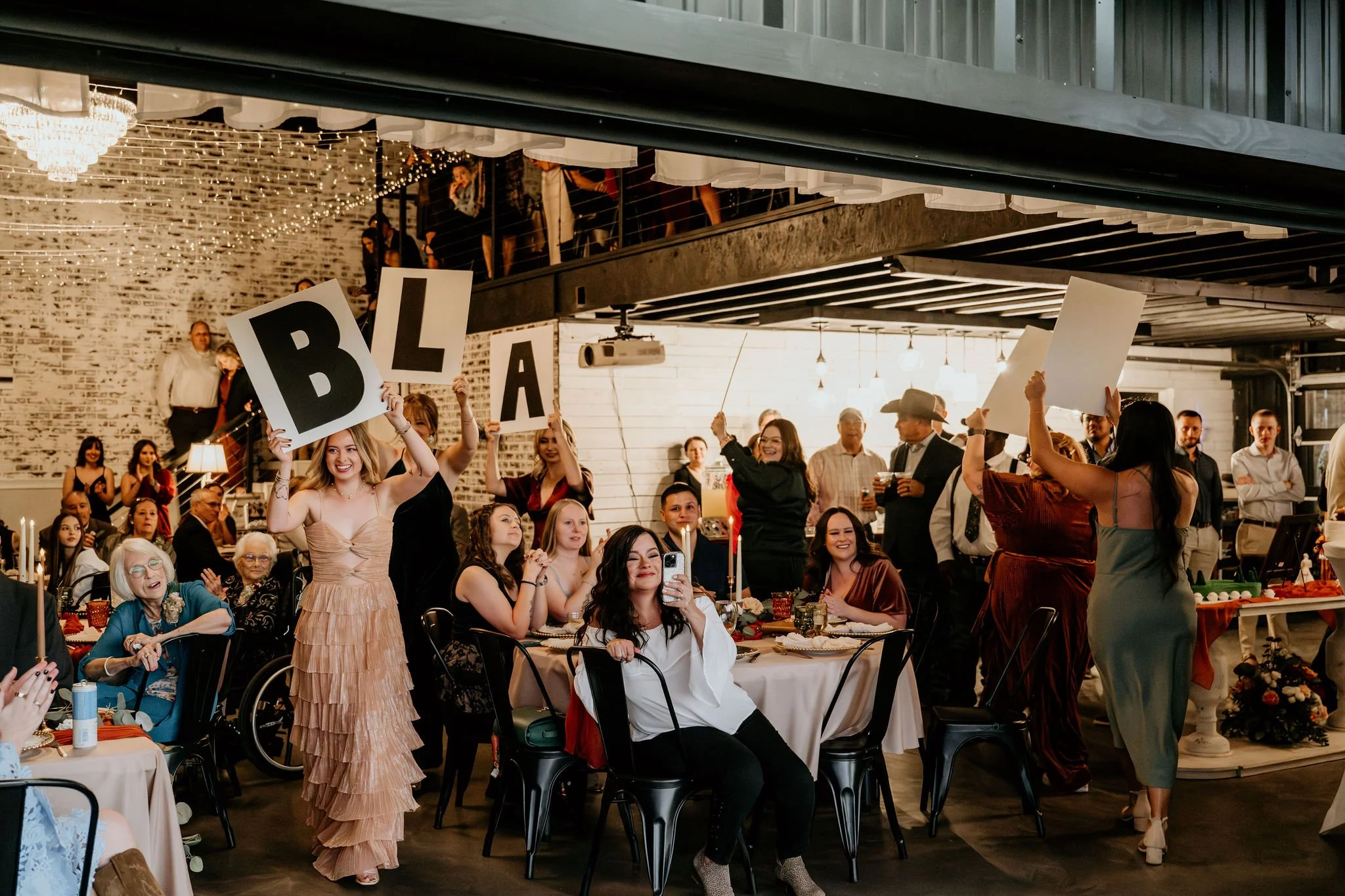 People at a celebration holding large letters that spell out "BLACK," with some seated at tables and others standing, some taking photos, in a decorated indoor venue.