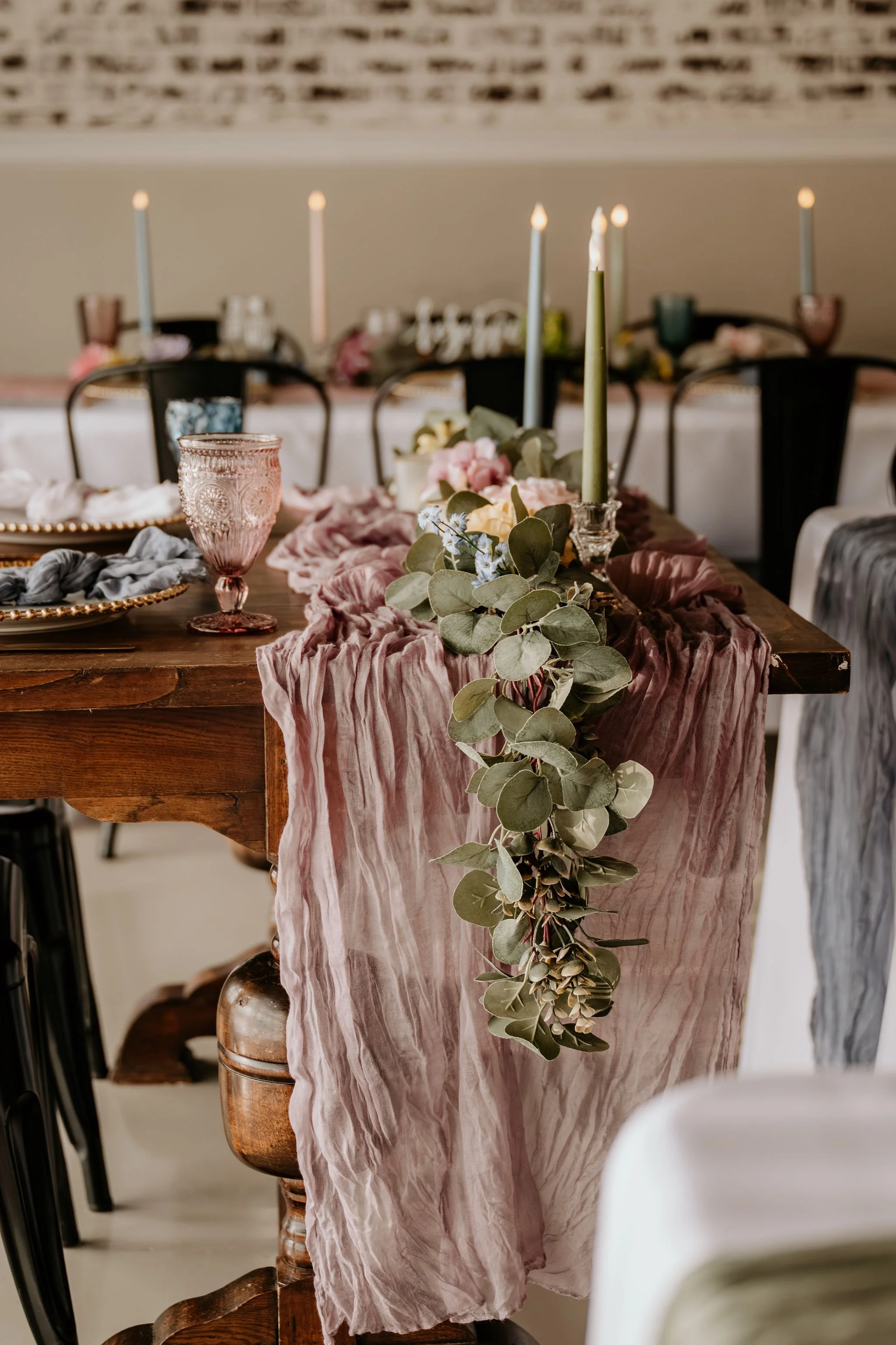 A decorated dining table with a pink fabric table runner, green candles, pink and blue flowers, and eucalyptus leaves cascading over the side.
