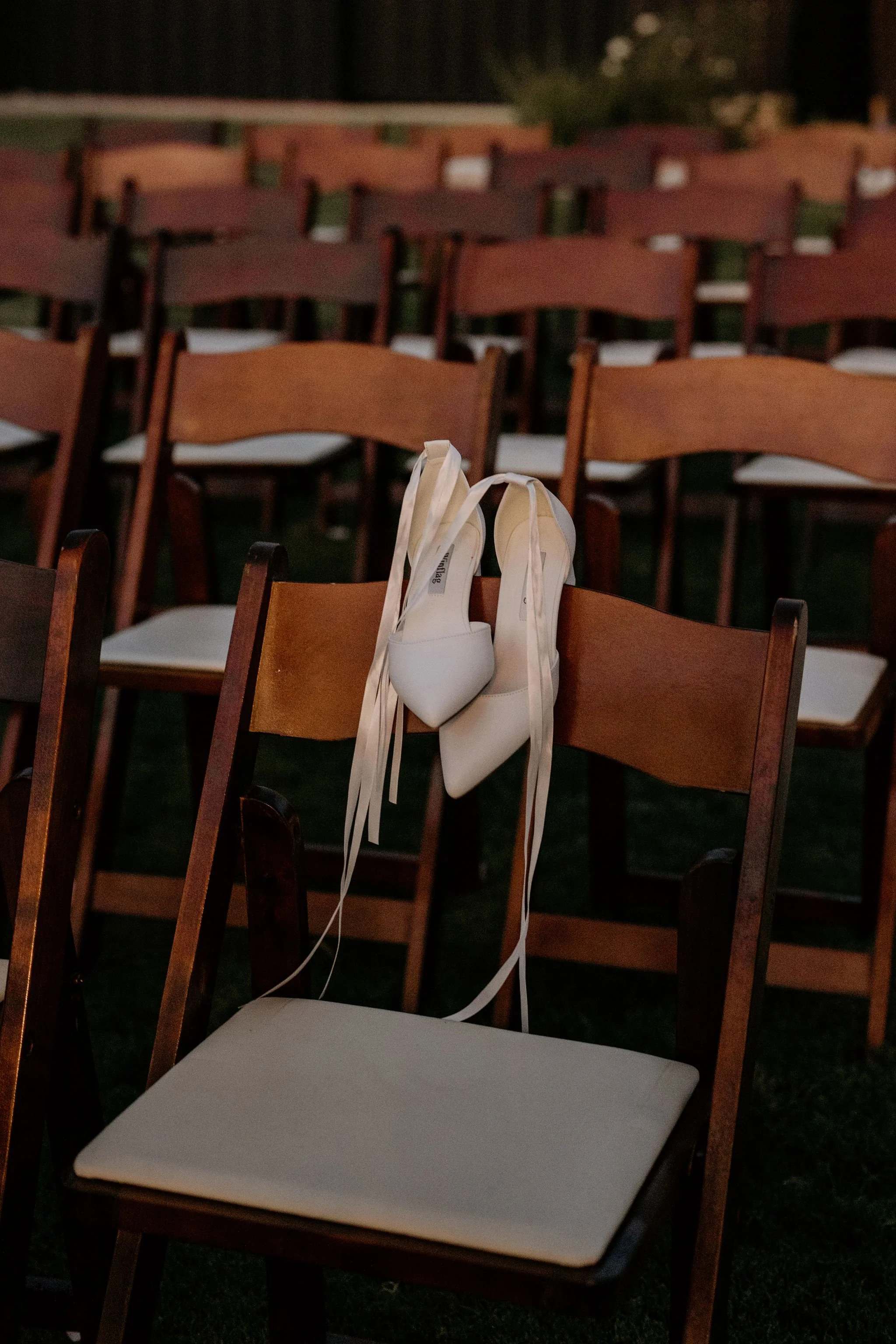 A wedding dress shoe hangs on a chair at an outdoor event with multiple rows of wooden chairs.
