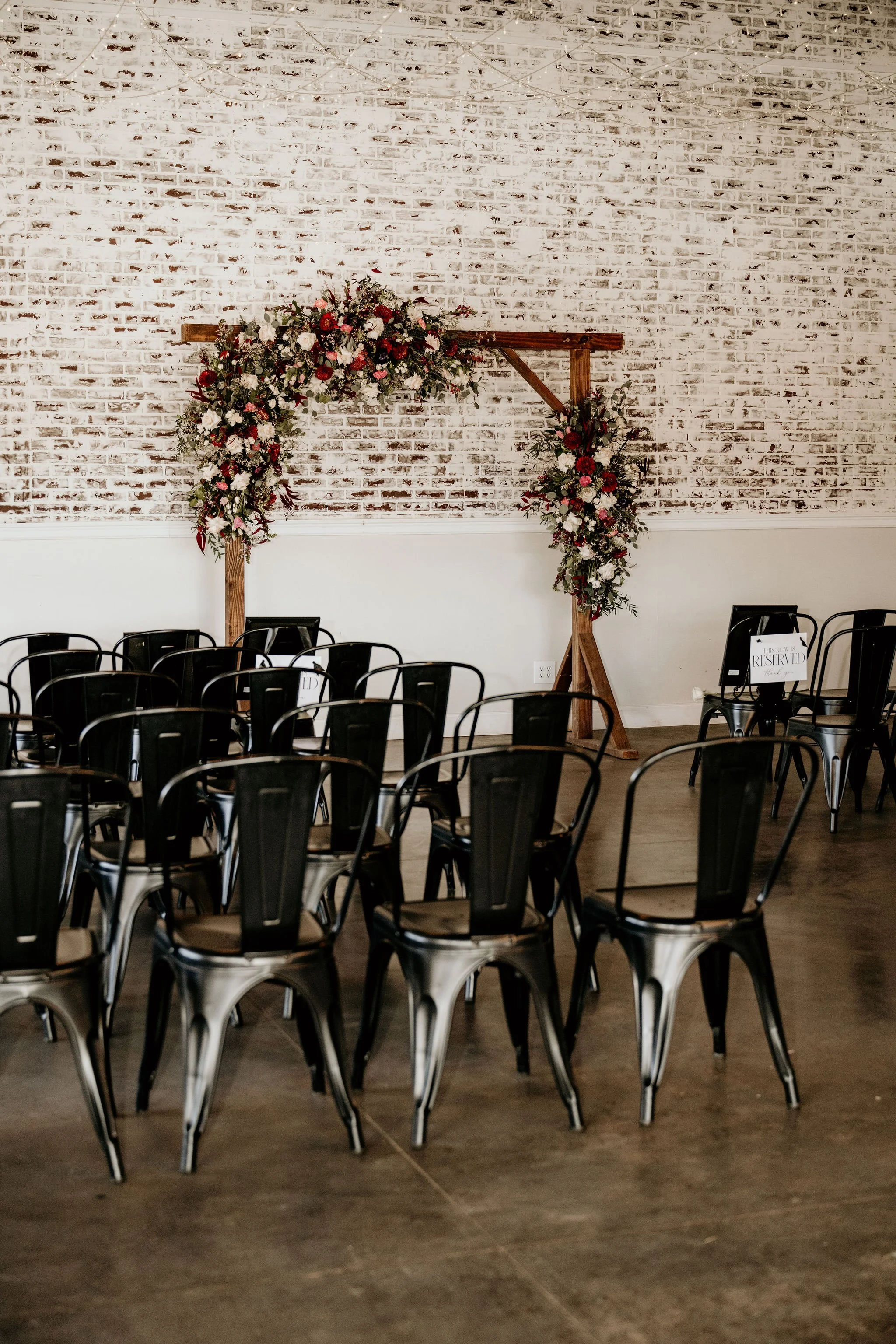 Wedding ceremony setup with black chairs arranged facing a floral arch on a brick wall.
