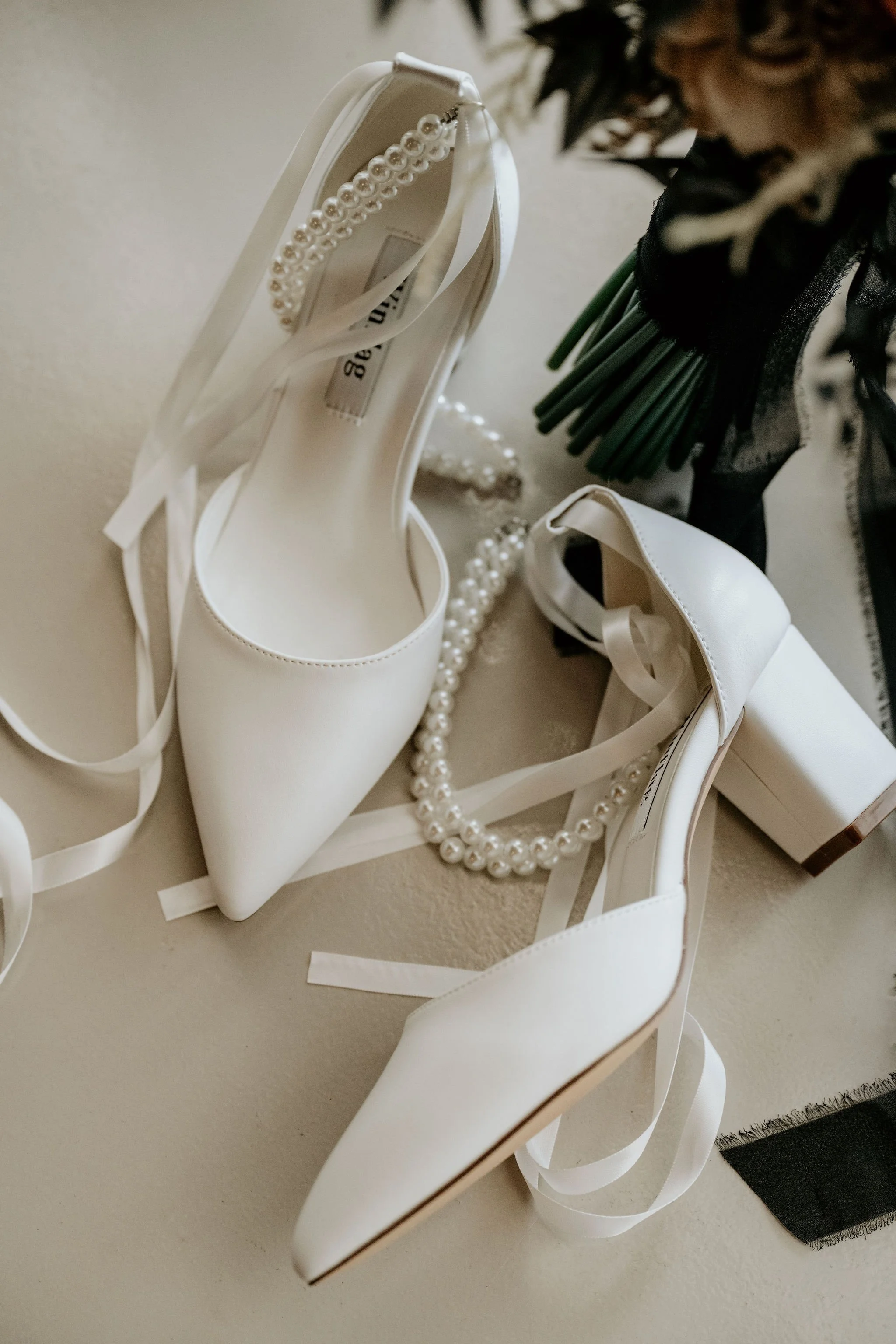 Pair of white high-heeled shoes with pointed toes, decorated with a pearl ankle strap, surrounded by pearl necklaces and floral arrangement.